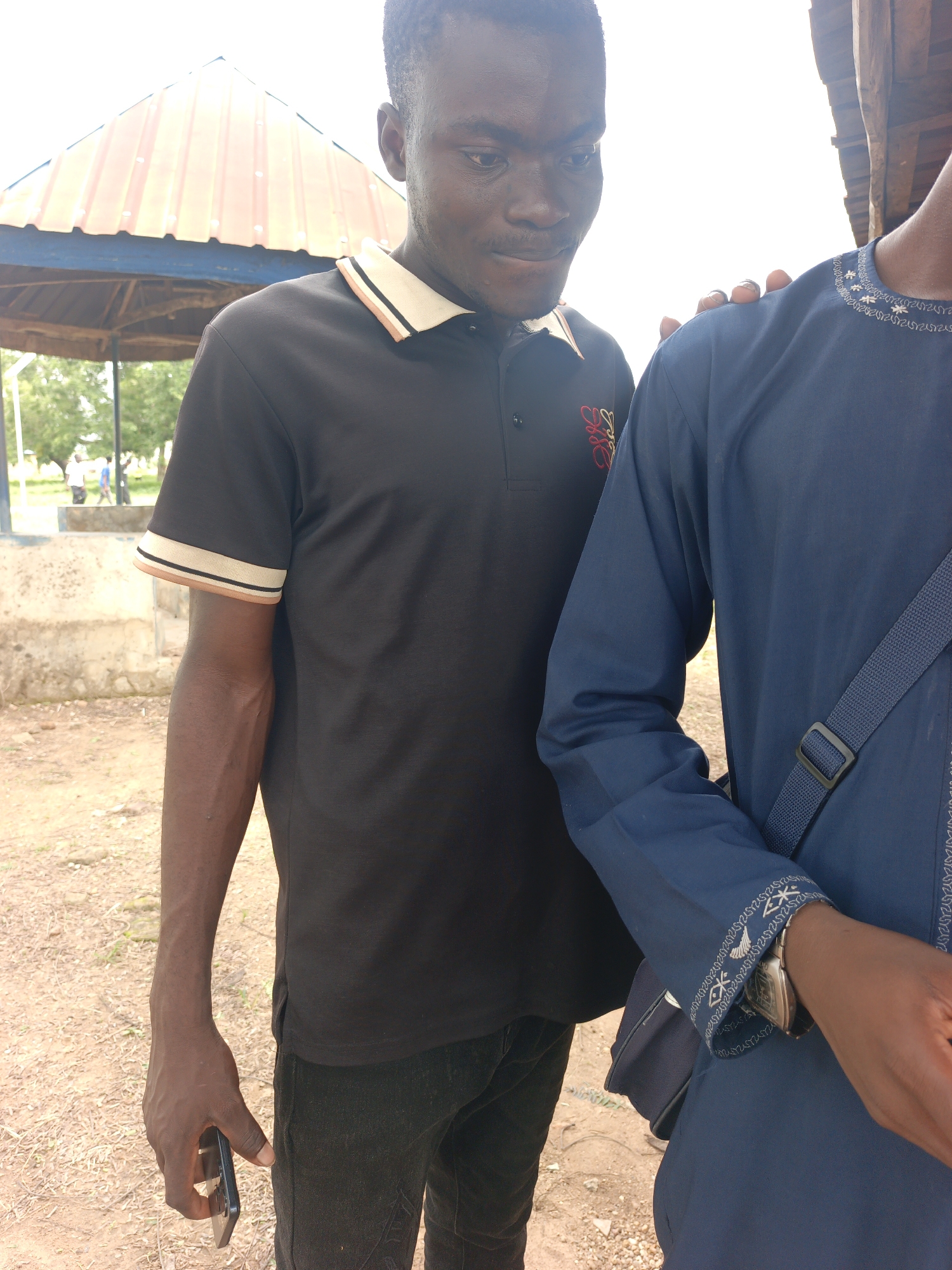 Two young men are standing outdoors. The man on the left is wearing a black polo shirt with striped trim on the collar and sleeves, and dark pants. He is holding a black smartphone in his left hand. The man on the right is wearing a blue robe with decorative white stitching on the cuffs and neckline. He has a dark blue strap across his chest. The background shows a sandy ground, a concrete wall, some trees, and a covered structure with a corrugated metal roof. The sky is bright and overcast.