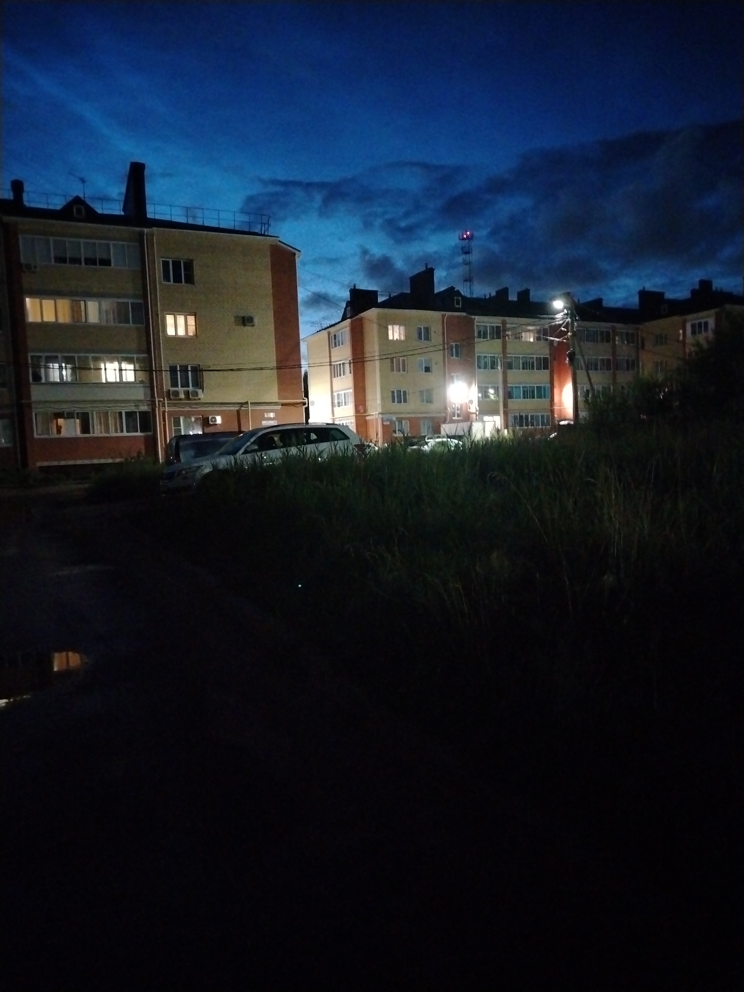 This is a nighttime, wide-angle photograph taken from a low vantage point, looking towards a multi-story apartment building. The sky is a deep, dark blue with wispy, illuminated clouds, suggesting twilight or dawn. In the foreground, tall, dark grass is visible, obscuring the lower parts of the scene. Two white cars are parked in front of the building, partially hidden by the grass. The apartment building itself is light yellow with reddish-brown accents and features numerous windows, many of which are illuminated with warm, interior light. A streetlamp is visible on a pole near the building, casting a bright glow. In the background, a cell tower with red and white lights is visible against the dark sky. The scene appears to be in a residential area of Yaroslavl, Russia, likely a courtyard or parking area adjacent to an apartment complex.