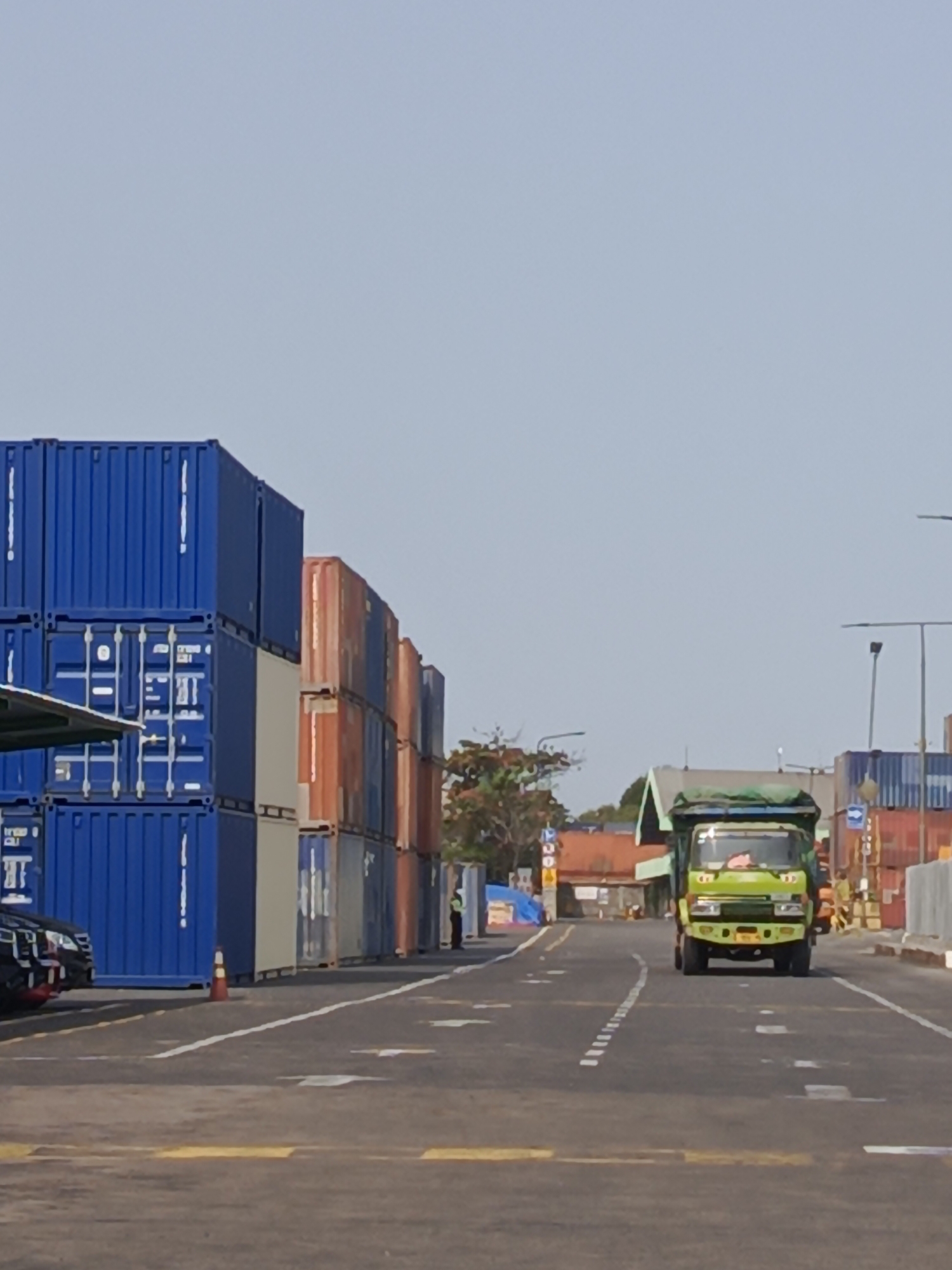 The image depicts a scene at a shipping container terminal, likely in the city of Surabaya, Indonesia. In the foreground, a green dump truck is driving down a paved road. The truck has a tarp covering its load and appears to be in motion. The road is marked with white and yellow lines.
To the left of the road, stacks of large blue shipping containers are visible, some reaching several levels high. To the right of the road, there are more stacks of containers, including some orange and grey ones. Beyond the containers, buildings with orange rooftops can be seen, suggesting an industrial or port area.
The sky is clear and light blue, indicating daytime. The overall environment appears to be an outdoor, operational port or logistics facility. A solitary figure, possibly a security guard or worker, is standing near the stacks of containers on the right side of the image, observing the scene. The presence of multiple shipping containers and a dump truck strongly suggests a location involved in the transport and storage of goods.