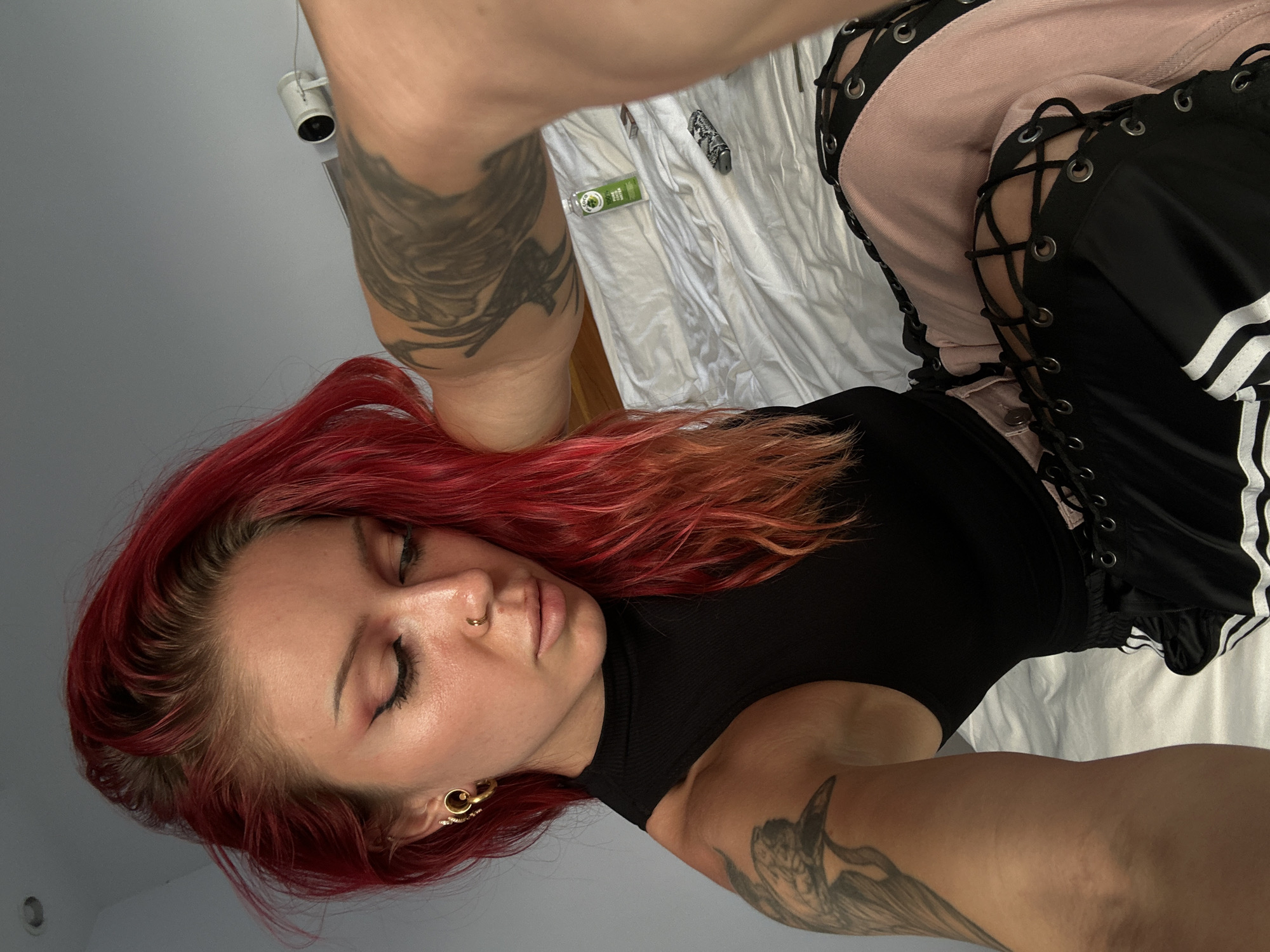 A woman with bright red hair is lying on a bed, taking a selfie. She is wearing a black sleeveless top and pink pants with black lacing details. She has tattoos on her arm and a nose ring. On the bed, there is a small bottle of what appears to be a drink, a lighter, and a small rectangular object. The room is sparsely decorated, with a grey wall in the background and a security camera mounted on the ceiling. The bed is covered in white sheets.