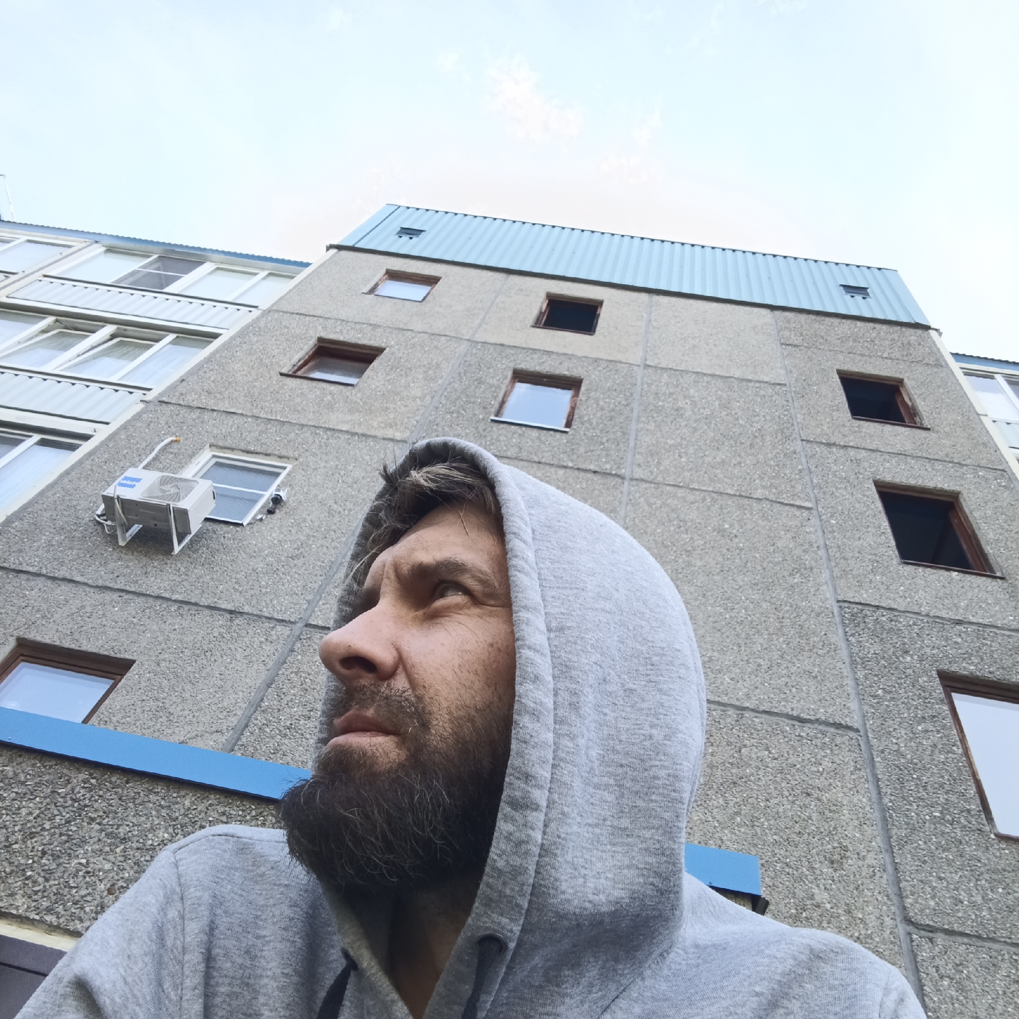 A man with a beard, wearing a grey hoodie, looks up at a tall, gray concrete building with multiple windows, located in Марковский, Russia. An air conditioning unit is mounted on one of the building's walls. The sky is visible above the building.
