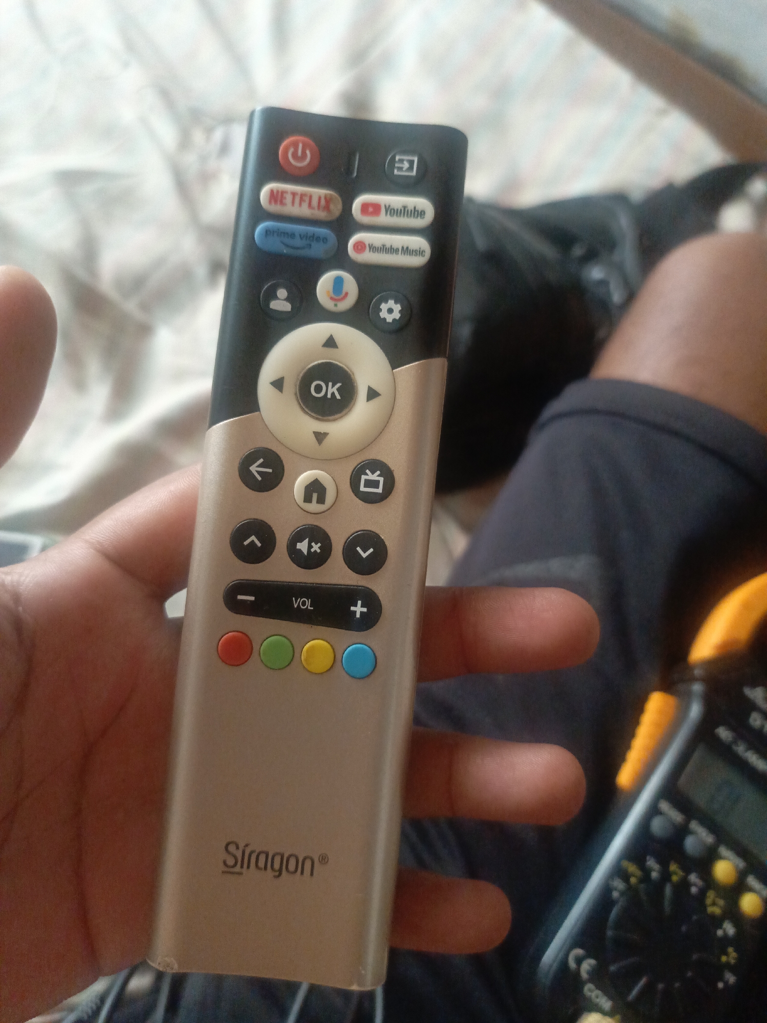 A hand holds a "Siragon" brand remote control. The remote is two-toned, black on top and champagne gold on the bottom. Prominent buttons include Netflix, YouTube, Prime Video, volume control, and a circular "OK" button surrounded by directional arrows. In the background, portions of a multimeter are visible, along with dark fabric, skin, and bedding with a light pattern. There is no information to suggest that the image was taken in Juan Griego, Venezuela.