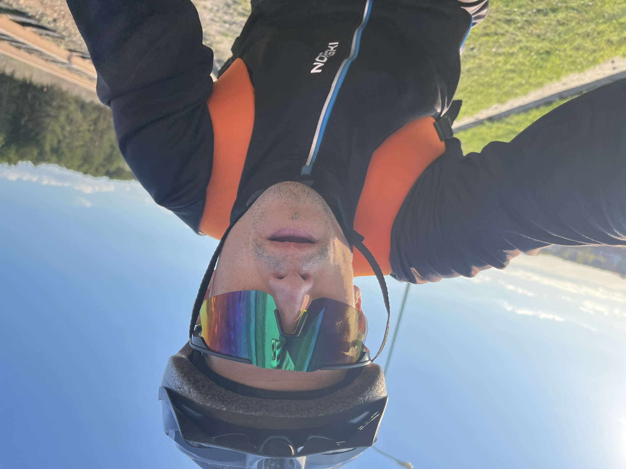 A person wearing a cycling helmet and iridescent sports sunglasses is facing the camera. The person is also wearing a black and orange jacket with a blue stripe and the word "NOSKI" on it. The background shows a blurred landscape with trees and what appears to be a river or a large body of water under a clear blue sky. The image is rotated 180 degrees. The location is Chaikovsky, Russia.