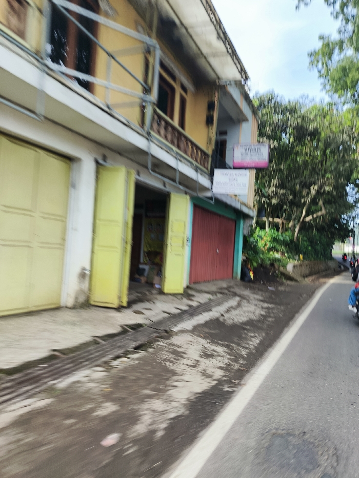 The image shows a street scene in Soreang, Indonesia. The photo was taken from a moving vehicle, making the image slightly blurry.  The street has a white dividing line in the center. The right side of the road shows the front of a building with a yellow facade. The building has a small balcony and a garage door with yellow horizontal panels. Next to the garage is a set of double doors. The doors are open and the inside of the building can be seen. The next door is red. The building is on the edge of a road that is paved with asphalt.  A small amount of green vegetation appears to be growing on a small embankment in the foreground. There are no people visible.  A motorcycle is shown traveling in the opposite direction to the viewer, but the driver cannot be seen. The photo is taken from a moving vehicle on a slightly curved, downhill section of road.   A sign is visible on the wall of the building. The sign is pink with black writing and appears to advertise "Warung Makan" (Indonesian for food stall) in the area.  The image is taken from a moving vehicle, resulting in motion blur. The photo is taken from a high angle, making the perspective seem wider.