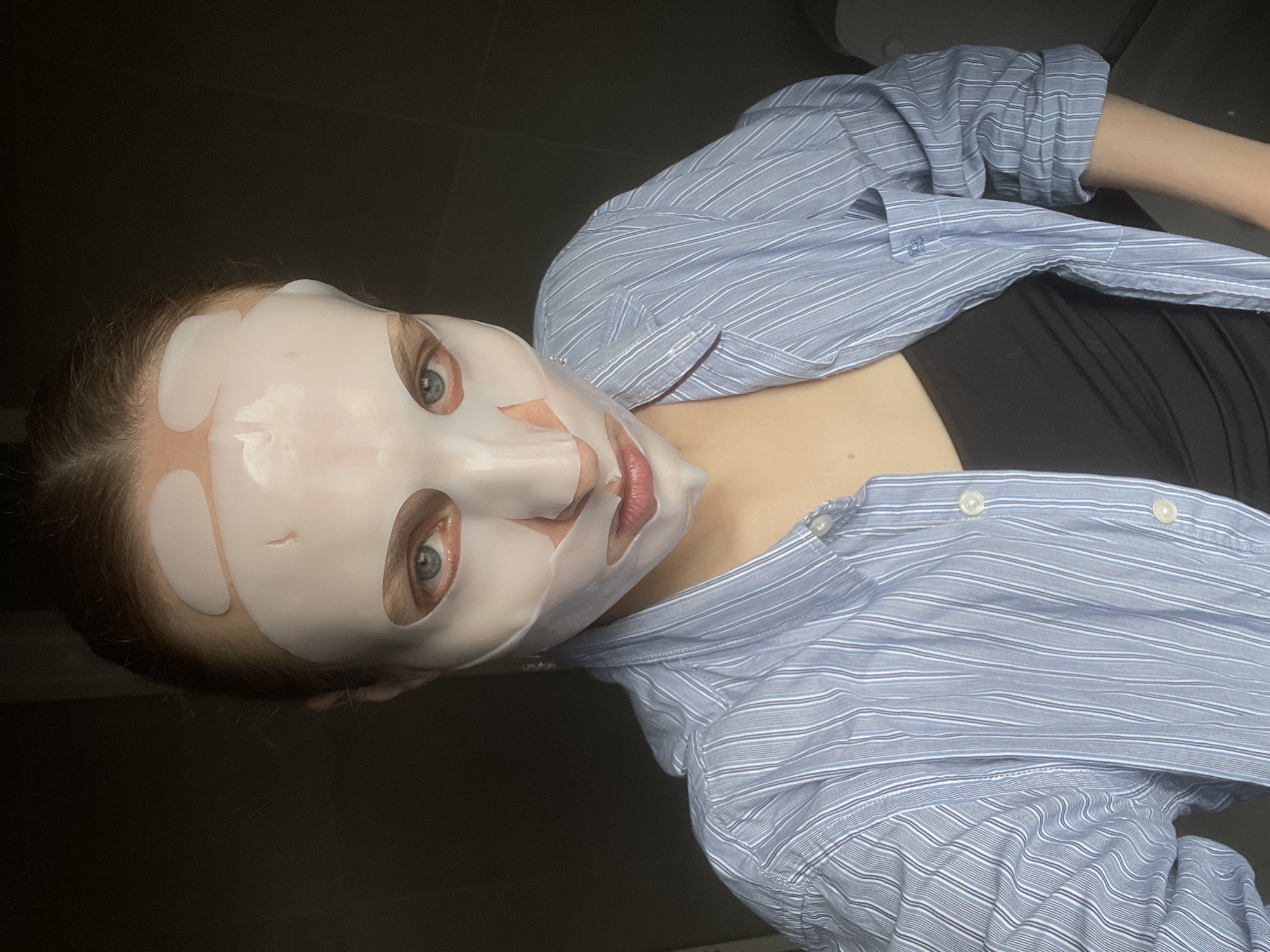 A woman with a facial mask is lying down wearing a blue and white striped collared shirt and a black top. The location is not specified in the image, and there is no indication it is in Подшивалово, Russia. The background appears to be a dark, solid color, possibly a wall. The woman is looking directly at the camera. The facial mask covers most of her face, with cutouts for her eyes and mouth. The mask appears to be a sheet mask, likely for skincare purposes.