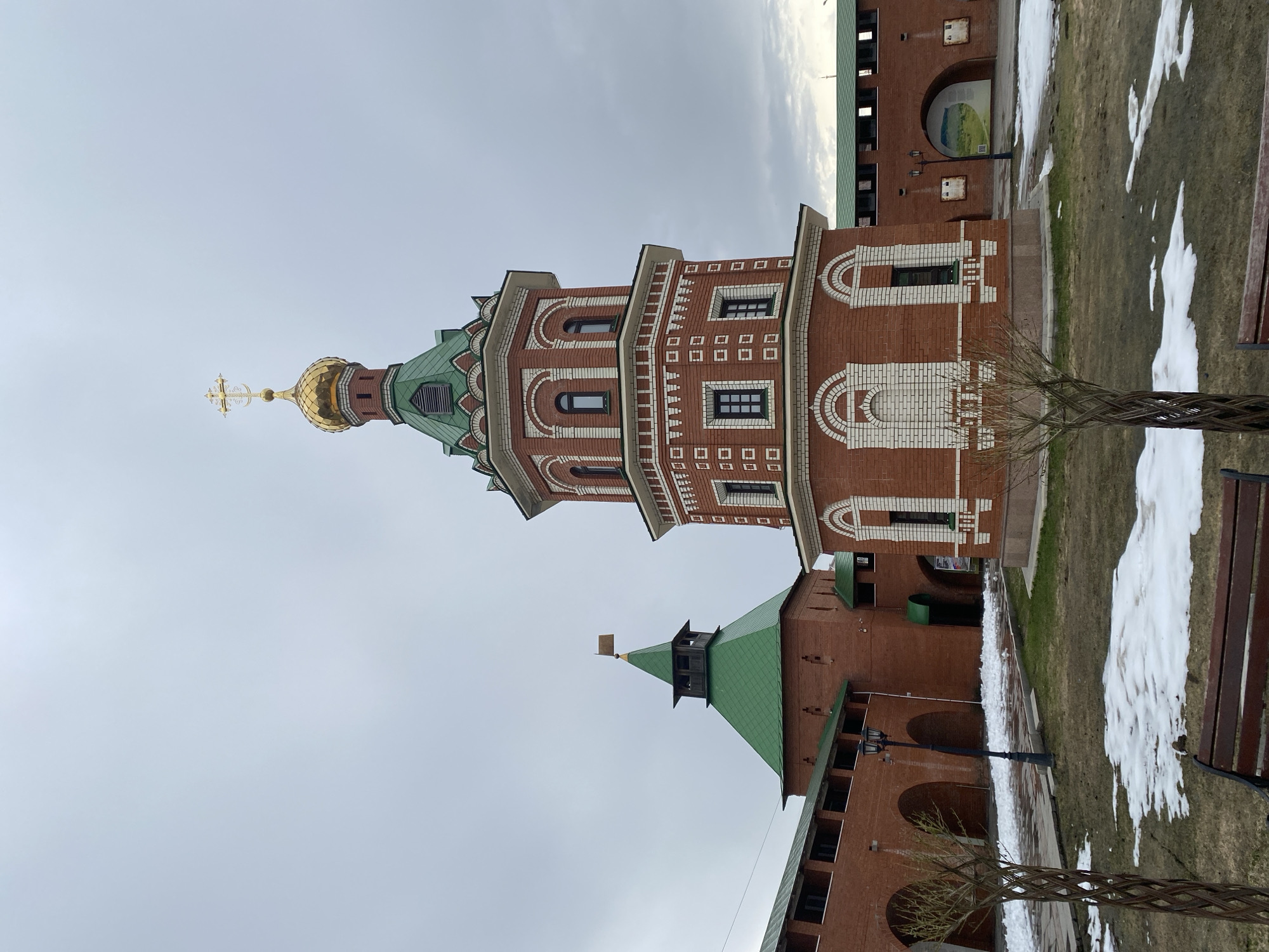 The image displays a daytime outdoor scene featuring distinctive Russian architecture under an overcast, light gray sky. The central subject is a tall, reddish-brown brick tower with a multi-tiered design, crowned by a golden onion-shaped dome topped with an Orthodox cross. The tower features a green, scale-patterned roof section, and its brickwork is adorned with intricate white decorative patterns, including arched window surrounds and geometric motifs.
Surrounding the main tower are other red brick buildings with green roofs, some featuring arched openings. One building has a large archway containing a visible sign with a landscape image. Another structure to the lower left has a pointed green roof with a small spire. The ground consists of grassy areas interspersed with paved pathways. Patches of melting snow are visible on the grass and along the paths, suggesting the season is late winter or early spring. A decorative, twisted sapling stands in the foreground, and a dark wooden park bench is visible on the right. A black lamppost is also present. The scene depicts a serene architectural complex, characteristic of Russian historical or religious sites, located in Yoshkar-Ola, Russia. No people are visible, and any text on signs is unreadable.