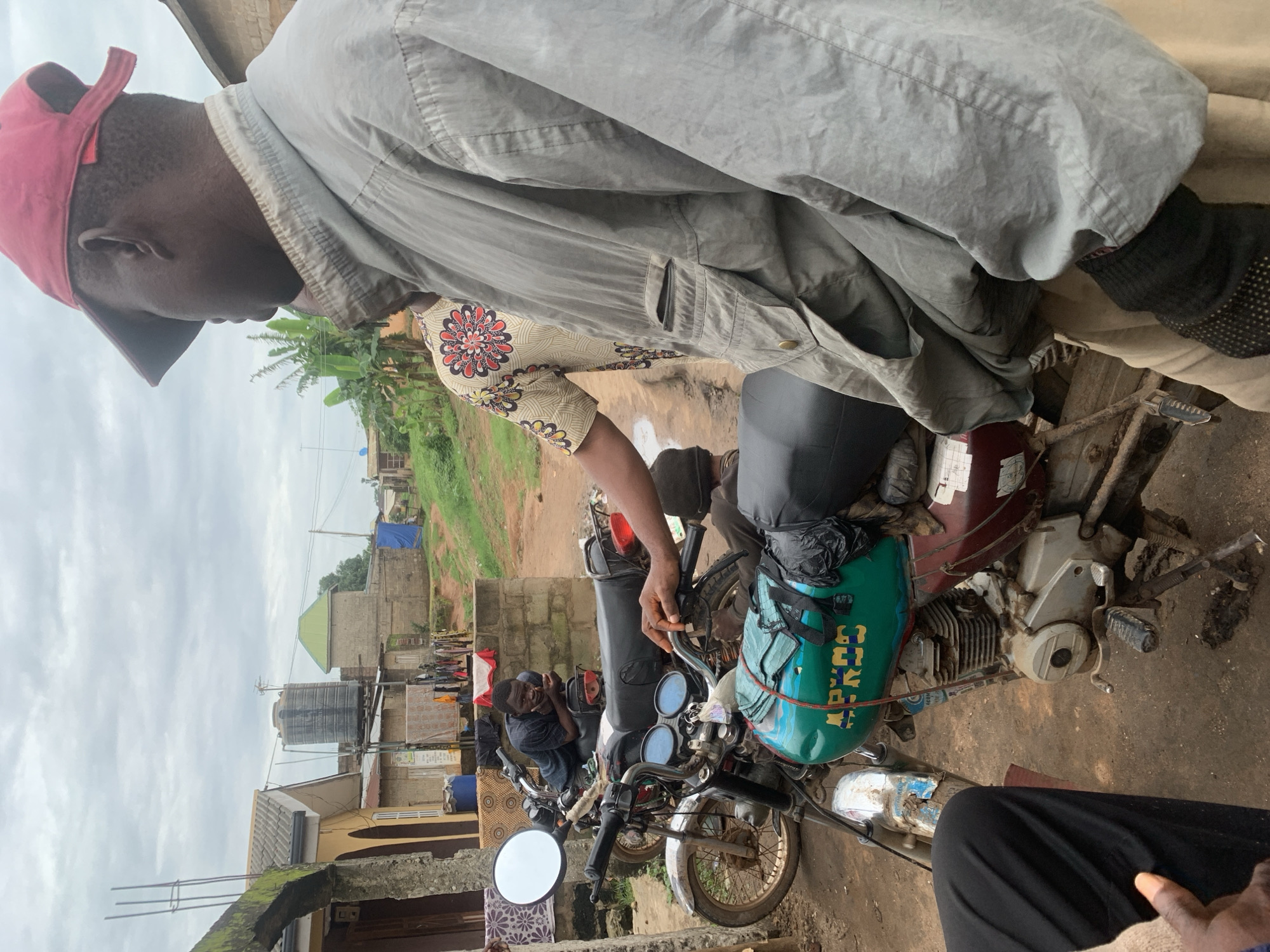 The image depicts a street scene in Shagamu, Nigeria, under an overcast sky. In the foreground, a person wearing a gray jacket and a red cap is seen from the shoulder up, looking to the right. To the left, another person in black pants is partially visible, with a hand extended forward.
The middle ground is dominated by two motorcycles. The motorcycle in the front is predominantly teal and maroon, with a black seat and green bags secured to it. The word "APROS" is written in yellow lettering on the teal portion of the fuel tank. The motorcycle behind it is black, with a black seat and a white circular mirror. A person is seen leaning on the handlebars of this second motorcycle, appearing to be looking down.
In the background, there are several buildings with corrugated metal roofs and brick walls. A large black water tank is visible on top of one of the structures. Clothes are hung out to dry on a line between two buildings. The ground is uneven and appears to be dirt and gravel. The overall environment suggests a residential or commercial area in a Nigerian city.