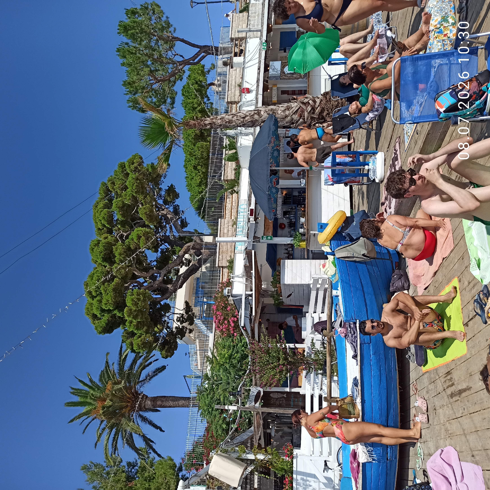 The image depicts a bustling beachfront scene in Trani, Italy, on a sunny day. A vibrant blue boat, likely used for decorative or functional purposes, rests on a wooden deck foreground. Several individuals are relaxing on towels and beach chairs. In the center, a woman in a multi-colored swimsuit stands near the blue boat, while a man with a beard and sunglasses sits on a lime green towel. To his right, another man with curly hair and sunglasses is seated on a towel. Further back, a woman in a red bikini sits on a towel. A beach bar with a counter, bottles of alcohol, and a dark umbrella is visible, with several people standing or being served. In the background, lush green trees, including palm trees, frame a white building with blue accents, suggesting a seaside establishment. The sky is a clear, bright blue. A green umbrella stands open near a woman in a blue bikini. The date stamp "08.07.2026 10:30" is superimposed on the lower right portion of the image.