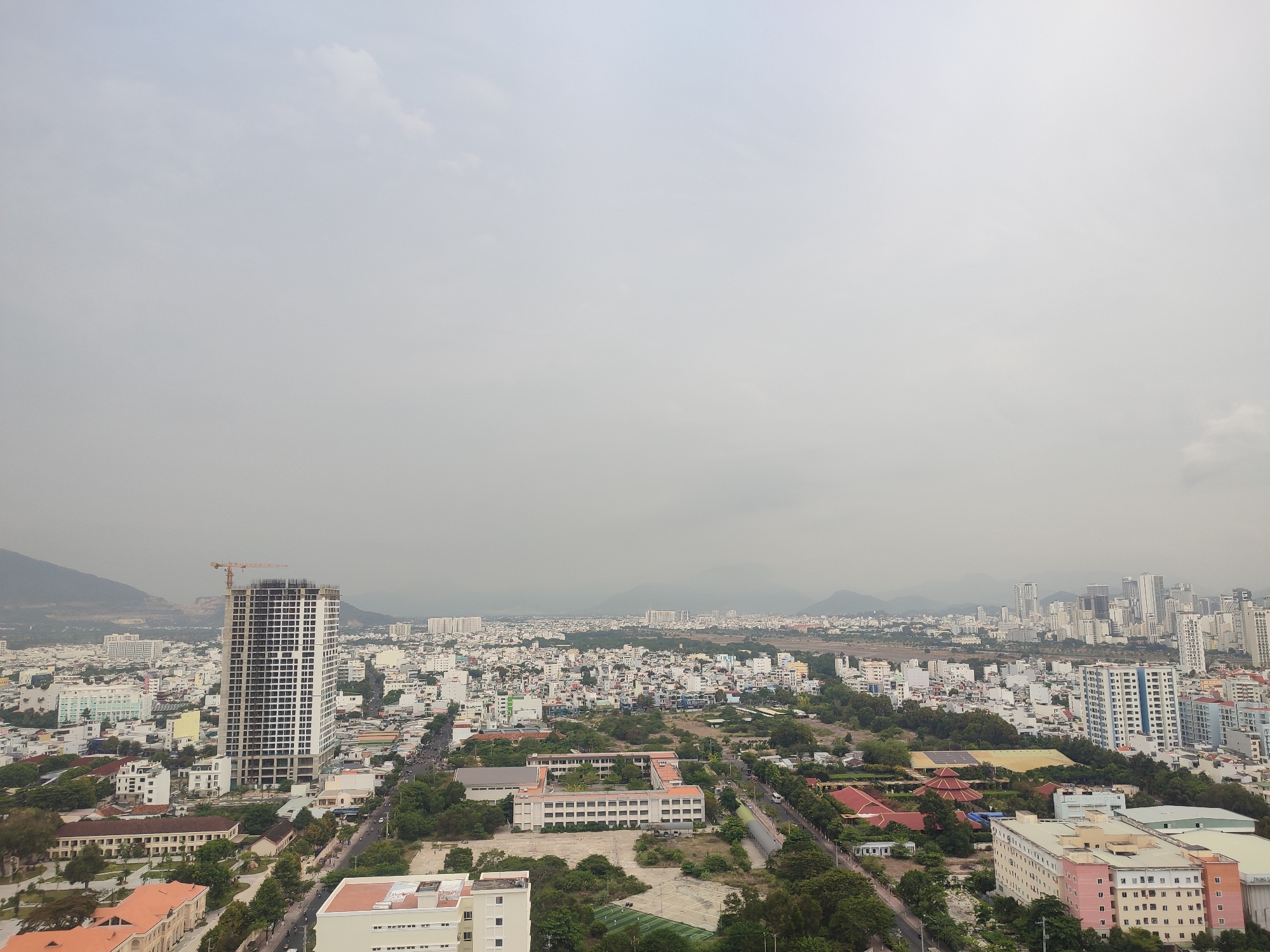 The image shows a wide-angle view of Nha Trang Ward, Vietnam, under an overcast sky. The scene is dominated by buildings of varying heights, from low-rise residential structures to tall skyscrapers, densely packed across the landscape. In the foreground are structures that appear to be schools, surrounded by trees. A large building under construction with a crane visible near the top is a focal point on the left side of the frame. In the distance, hazy mountains rise above the cityscape. The overall impression is a densely populated urban area with a mix of old and new architecture, and industrial activity under a dull sky.