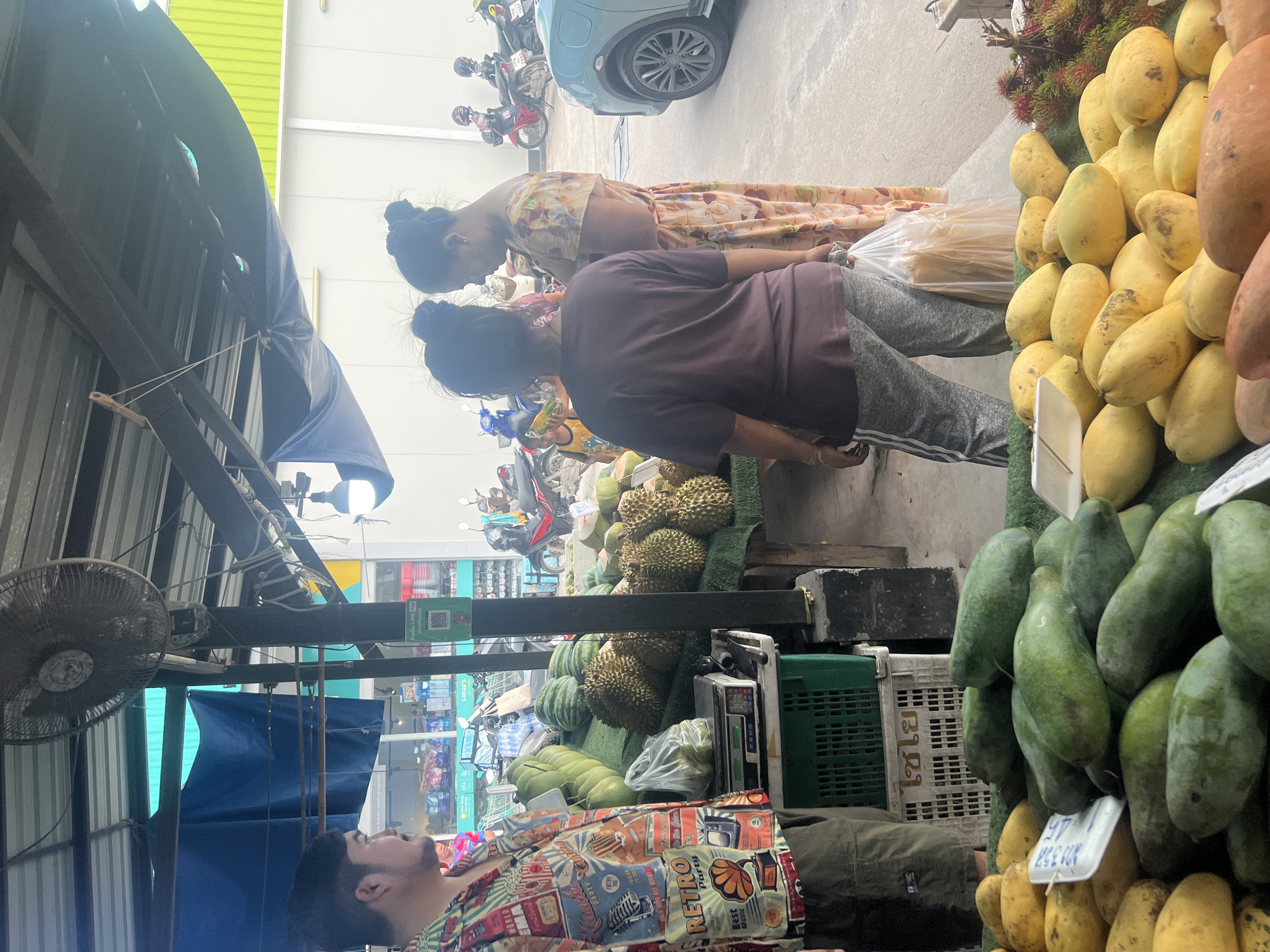In a street market in the Ko Samui District of Thailand, a man wearing a patterned shirt stands by a stall piled high with fruits. In front of him is a digital scale on top of a green plastic crate, and beside the scale, a plastic bag filled with what appears to be green fruit. Behind the man, two women are engaged in conversation while standing next to a display of durians and other fruits. The market is outdoors, under a tin roof supported by metal poles, with a blue tarp hanging from the roof. In the background, several motorcycles are parked, and a light blue car is visible. The ground is paved with concrete, and a white building with green trim is behind the market stalls. The overall scene depicts a bustling fruit market, likely during daylight hours.