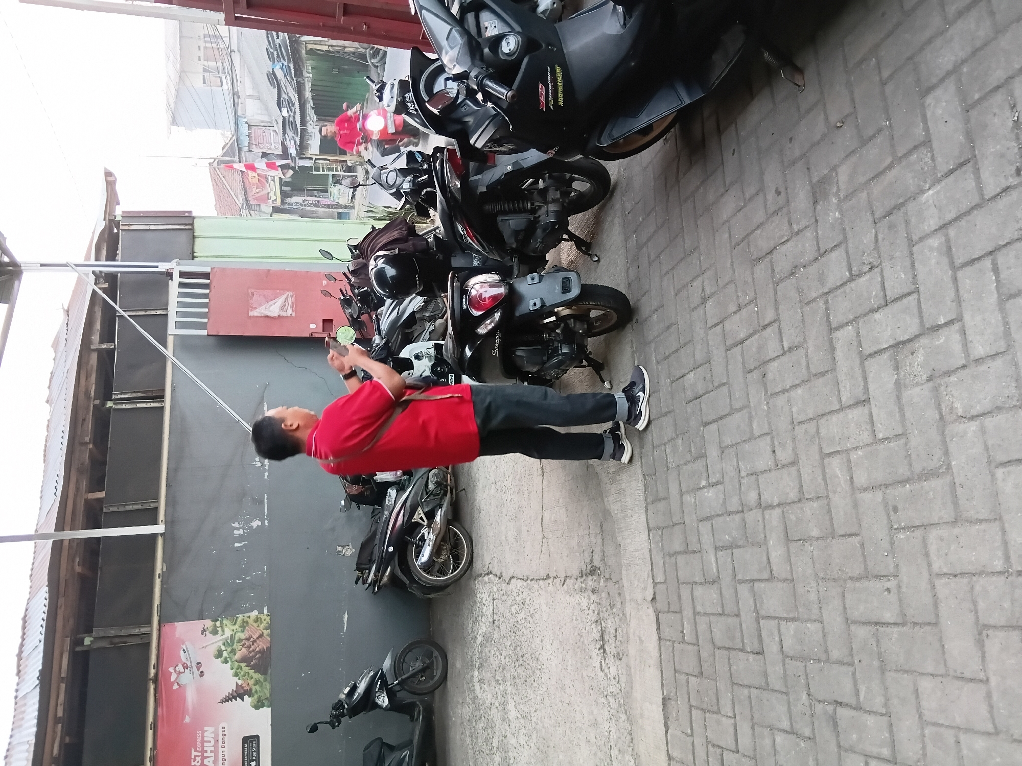 In an outdoor urban setting, likely a parking area in Bekasi, Indonesia, a man wearing a red polo shirt and dark pants stands with a cell phone to his ear, appearing to be engaged in a conversation. He is positioned amidst a cluster of parked motorcycles, mostly dark-colored scooters. One motorcycle in the foreground has a prominent "X-Ride" sticker on its side. Further back, another man in a red shirt is riding a scooter, illuminated by its headlights. The background reveals a row of buildings and structures, some with corrugated metal roofs, indicative of a commercial or residential area. The ground is paved with interlocking grey bricks. A large mural advertising a service, possibly for deliveries, is visible on the grey wall behind the man. The scene suggests a typical streetscape in Indonesia.