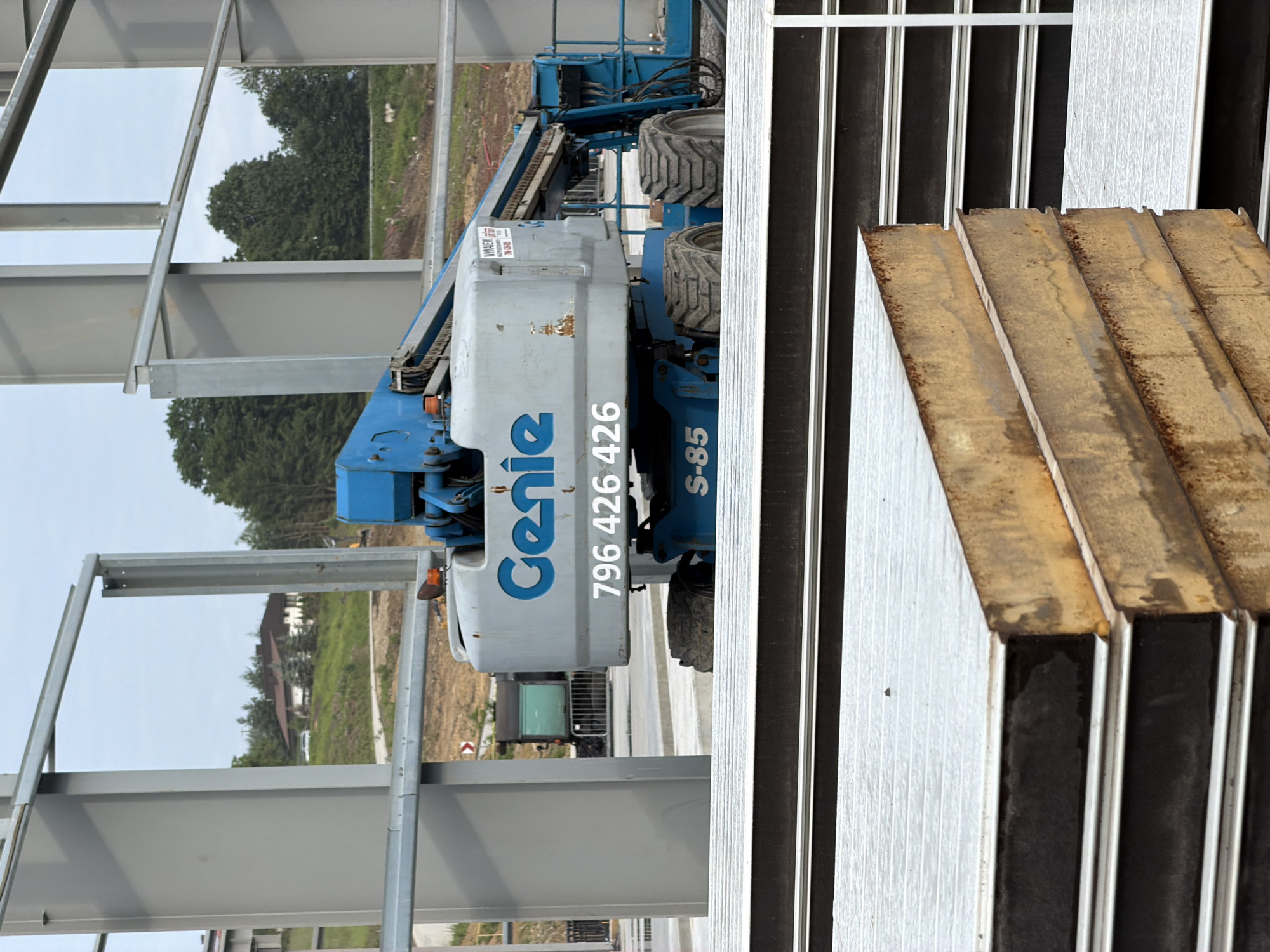 A blue and gray Genie aerial work platform is parked on a construction site in Stróża, Poland. The platform is positioned in front of a building under construction, characterized by its large gray metal beams. In the background, a green hill dotted with trees and a house can be seen. In the foreground, several large, flat, insulated panels are stacked, with some showing signs of rust. The sky is overcast. No people are visible in the image.