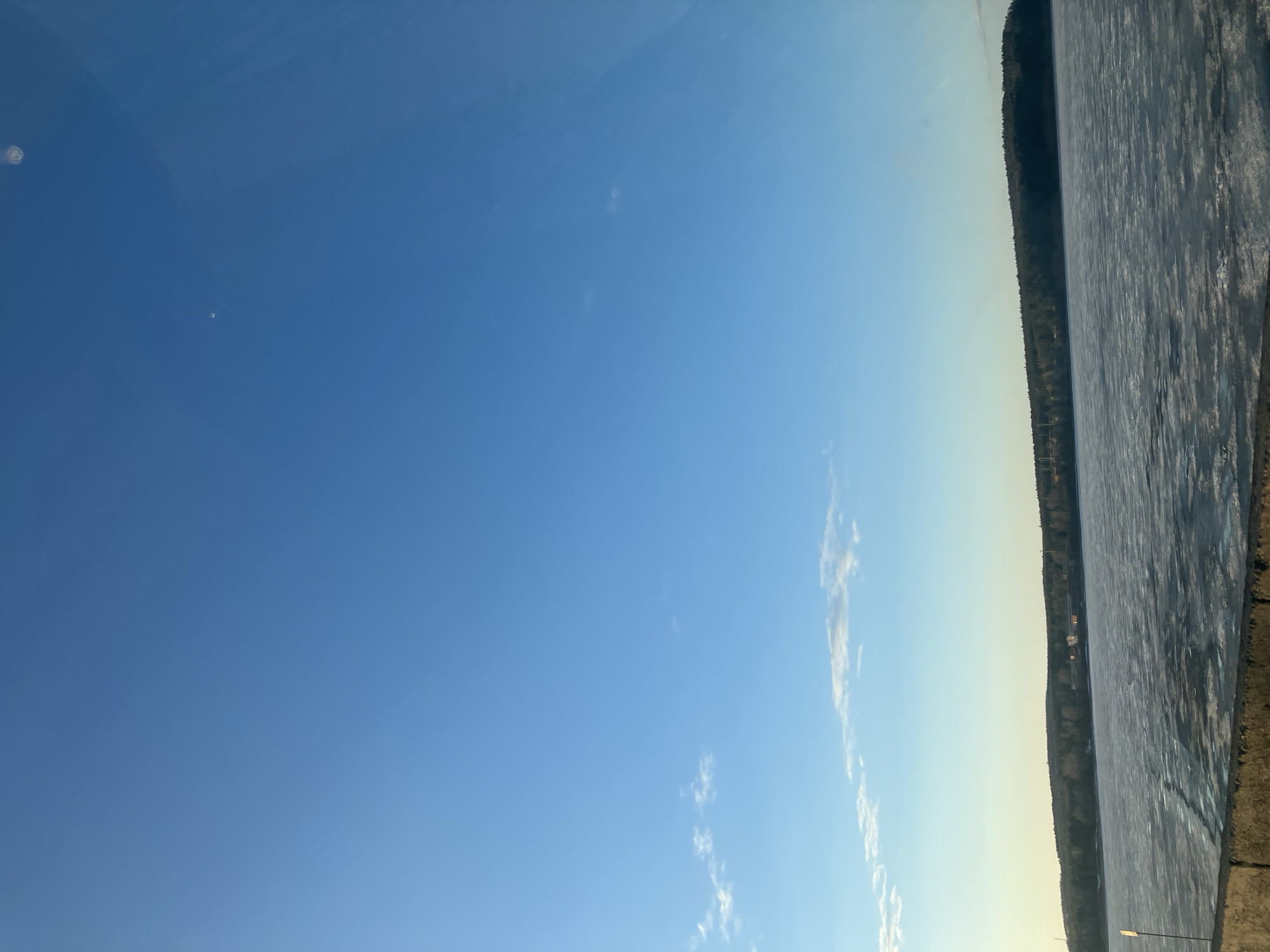 The image depicts a landscape in the city of Chaikovsky, Russia. The sky is a clear blue with a few scattered clouds. On the right side of the image, there is a frozen lake or body of water, with visible ice patterns. A treeline runs along the edge of the water. In the middle ground, buildings or structures are barely visible behind the treeline. The foreground consists of a strip of land or shore, appearing dry. The photo seems to have been taken during the day in sunny conditions.