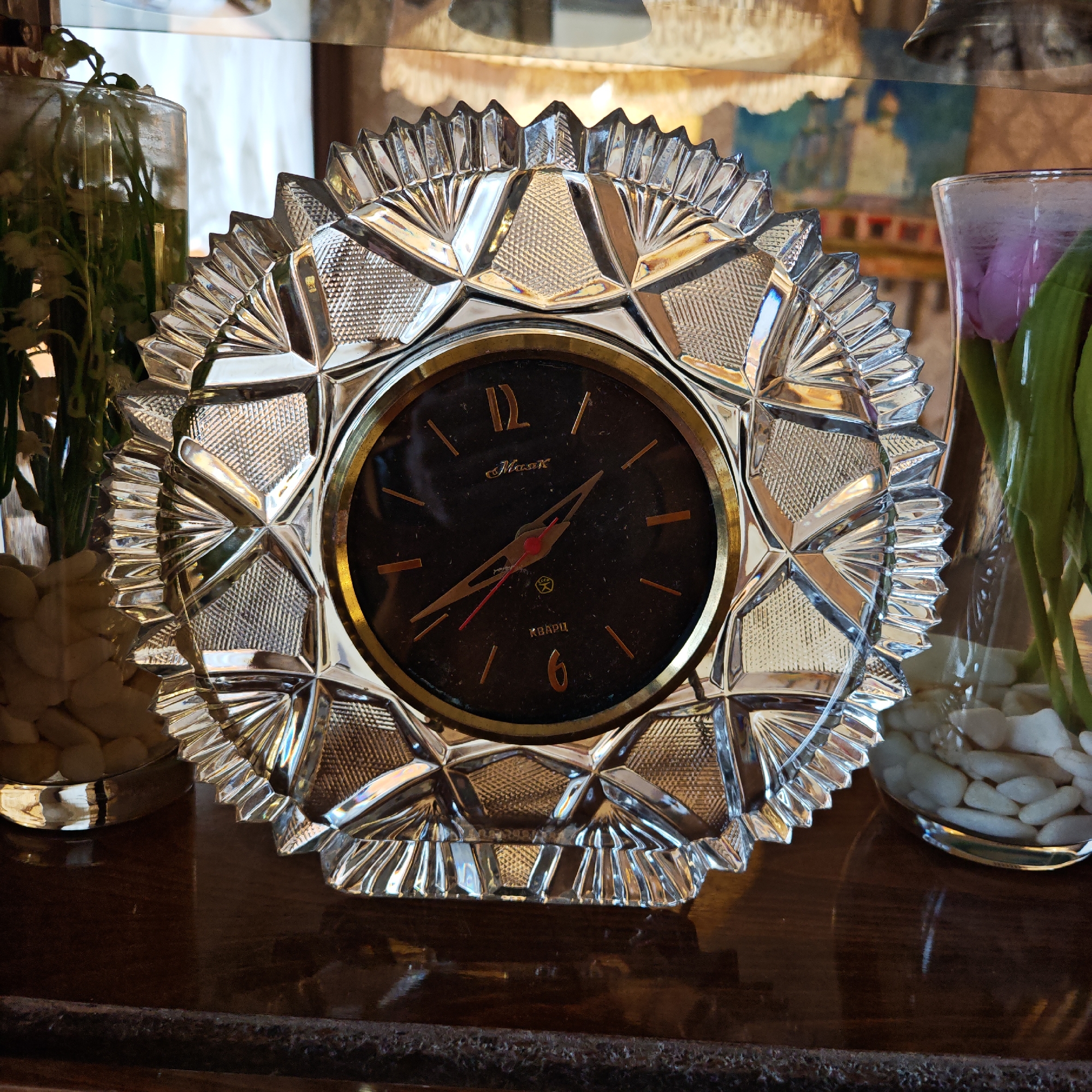 A crystal clock sits on a wooden surface, flanked by two glass vases containing plants and white stones. The clock has a black face with gold numerals and hands. In the top of the clockface, the name "Mayak" is written in Cyrillic script. Beneath, there is the word "Quartz" written in Cyrillic script. The crystal casing of the clock is faceted. The vases on either side contain green foliage, and one contains a purple flower bud. The scene is indoors, likely a home interior. It is not possible to determine the location in Rostov, Russia from this image alone.