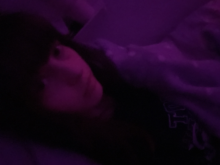 A person with dark hair is shown in low lighting with a purple hue. The individual's face is partially visible, tilted towards the camera, and appears to be resting on something. The person is wearing a dark garment that has visible lettering. The rest of the background is obscured, but it seems to be a soft, textured surface, possibly bedding or a blanket.