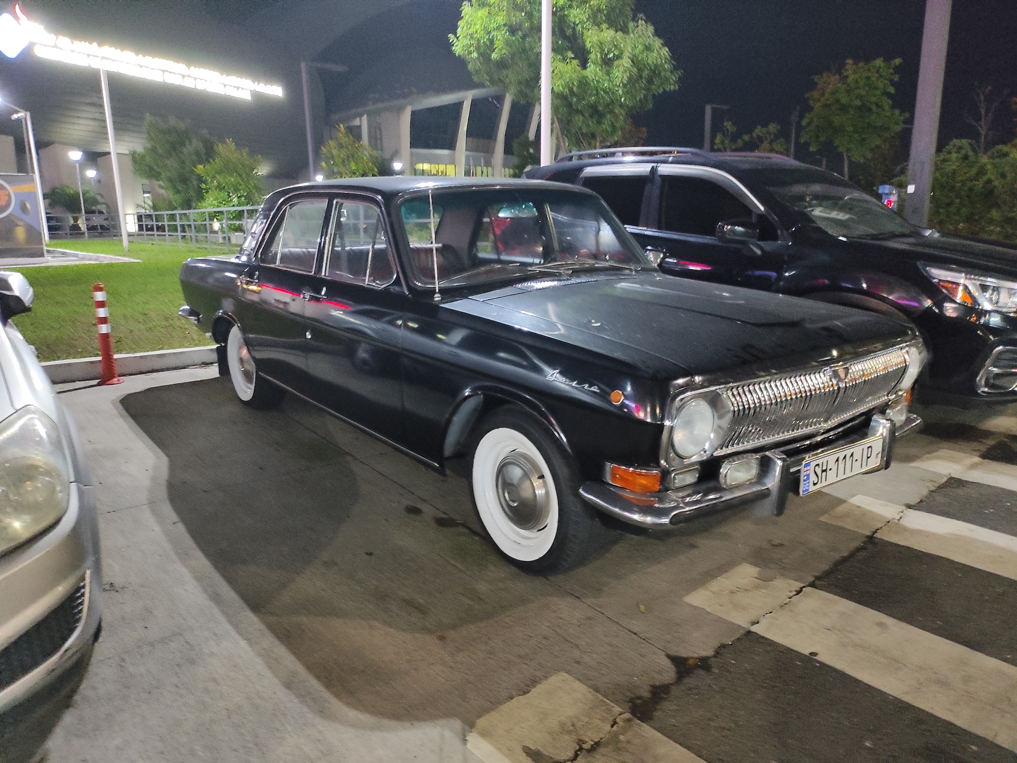 A black, vintage sedan with white-walled tires is parked on a street at night. The car has a chrome grille and bumper, and a license plate that reads "SH-111-IP". A modern black SUV is parked behind the sedan, and part of a silver car is visible to the left of the sedan. The background shows a building with lights and trees. The scene appears to be a parking lot or street in a city.