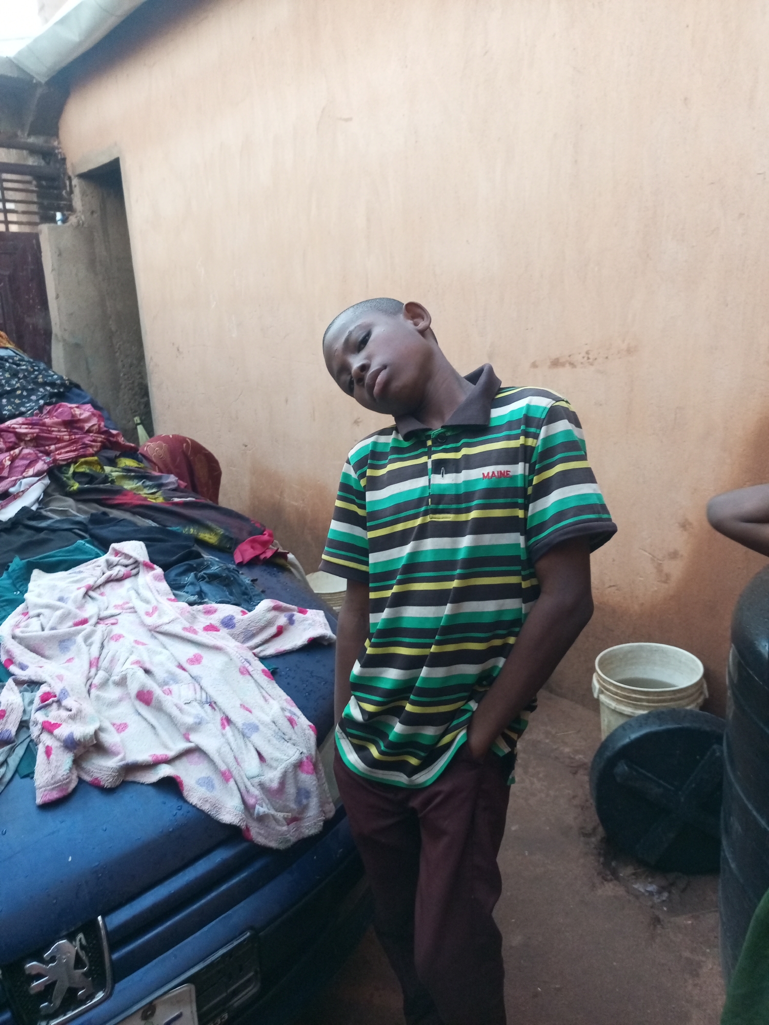 In a dusty, outdoor setting that appears to be the city of Tempure, Nigeria, a young boy stands with his head tilted back and one hand in his pocket. He is wearing a horizontally striped polo shirt with bands of green, black, yellow, and white, and the word "MAINE" is visible in red lettering on the chest. His pants are a dark, reddish-brown. To his left, a pile of colorful clothing, including patterned fabrics and a pink and white blanket with hearts, is draped over what appears to be the hood of a dark blue car, identifiable by a silver Peugeot emblem. In the background, a light brown wall forms the backdrop. To the boy's right, a white bucket filled with murky water and a black plastic lid are visible on the ground, alongside a large black water tank. The ground is composed of reddish-brown dirt.