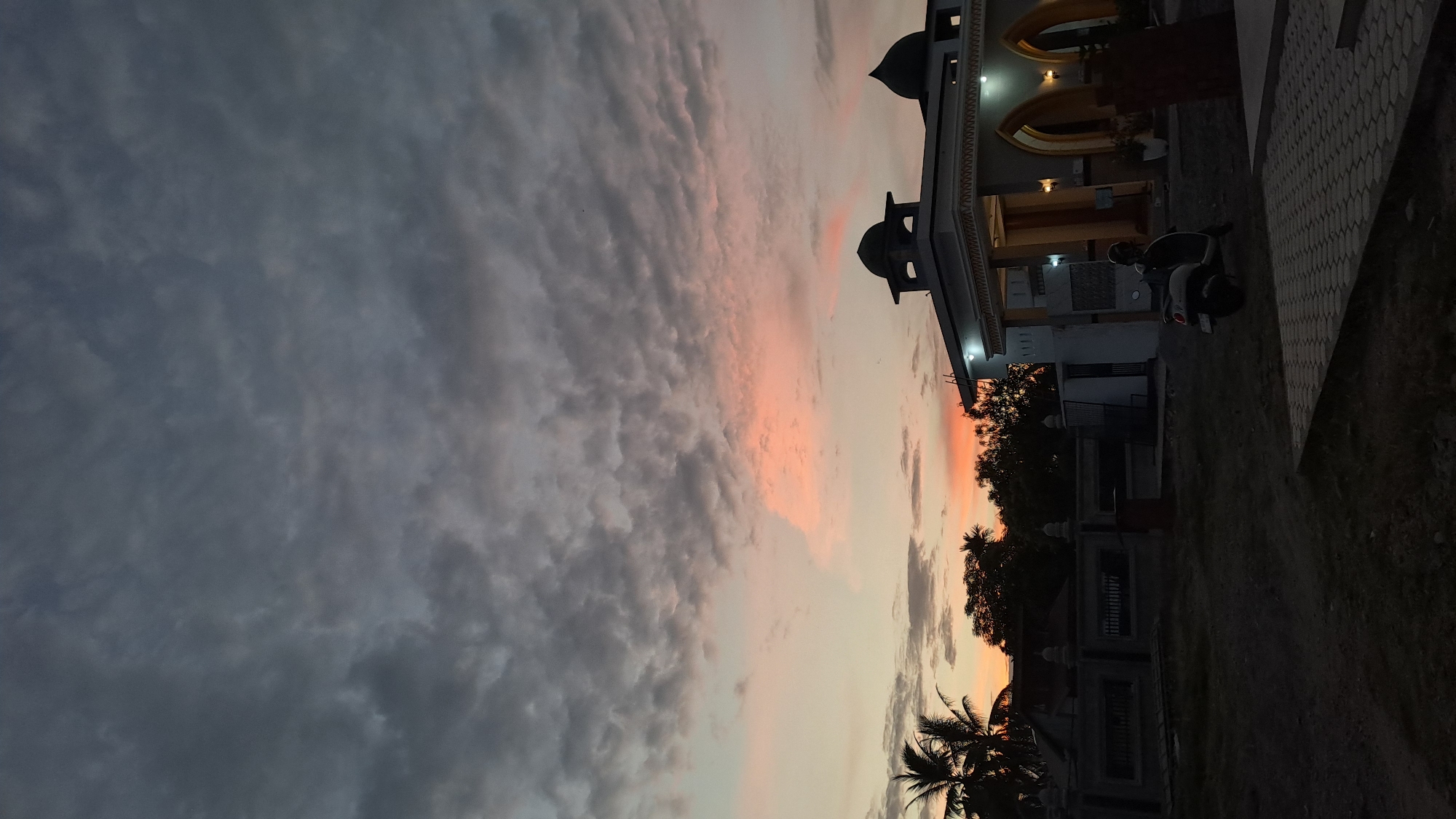 A mosque with two domes is visible against a cloudy sky at sunset. The sky has patches of pink and orange. In front of the mosque, a scooter is parked. Palm trees and other foliage are present in the background. A paved area is in the foreground. The scene appears to be in a city or town, possibly Lhokawe Teungoh, Indonesia.