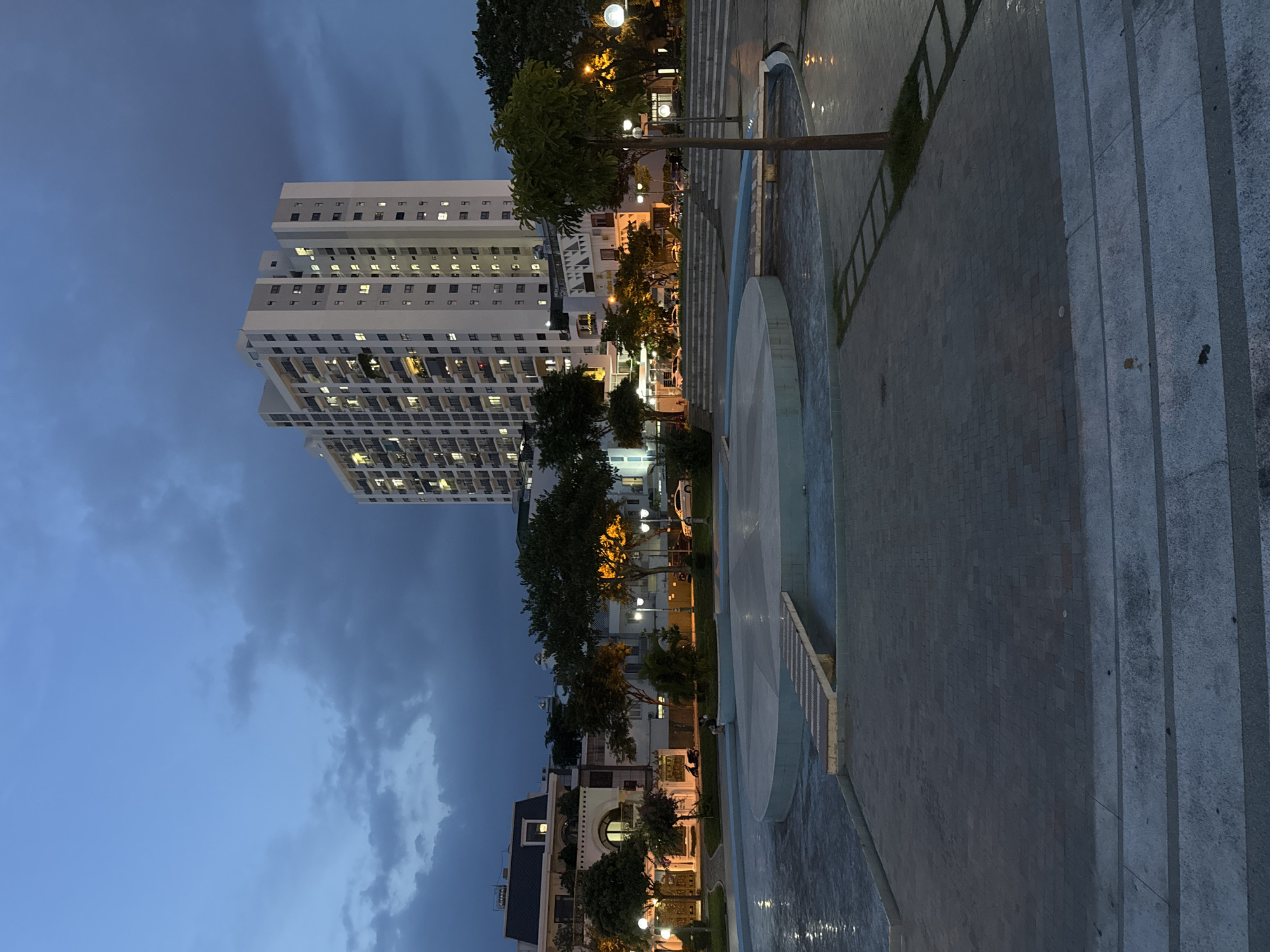 The image depicts an outdoor scene at dusk in Nam Nha Trang Ward, Vietnam. In the background, a tall, white apartment building with numerous illuminated windows dominates the skyline. In the middle ground, several smaller buildings with residential or commercial facades are visible, interspersed with trees. A multi-tiered water feature or fountain occupies the foreground, with a paved area surrounding it. The sky is a deep blue, with scattered clouds, indicating the transition from day to night. Streetlights and lights from the buildings cast a warm glow on the scene. Paved walkways and stairs are evident, suggesting a public space such as a park or plaza. A single tree stands prominently in the right foreground. No specific actions or interactions between people are clearly discernible, though there are hints of individuals in the distance. The overall impression is one of urban evening ambiance.