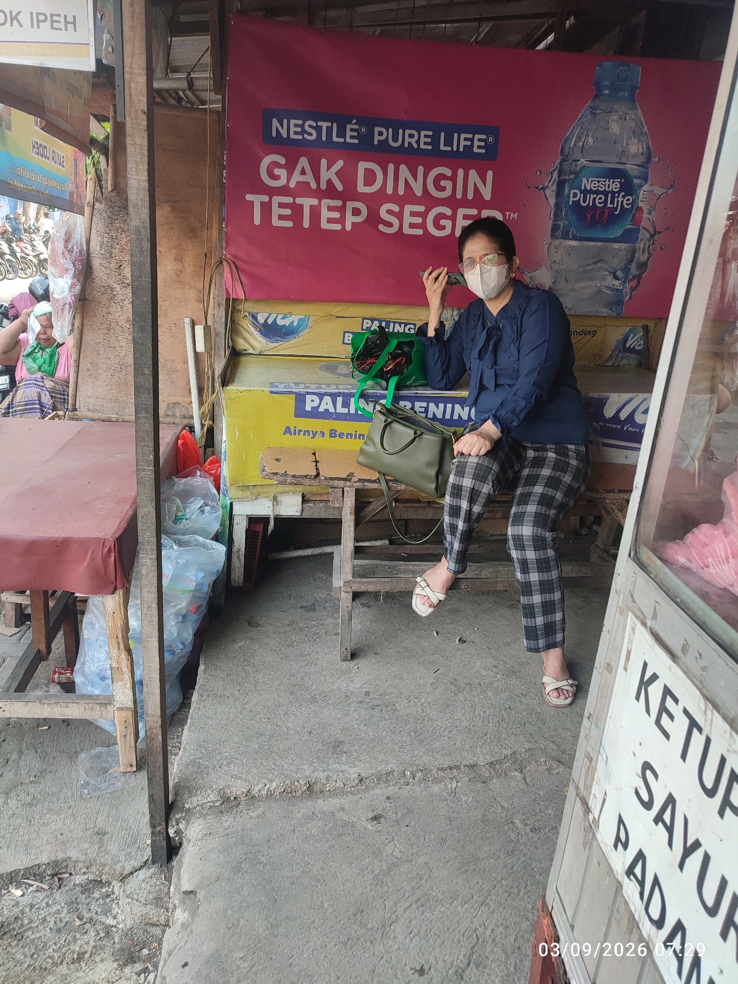 The image shows a street-level view of a small market stall in East Jakarta, Indonesia. A woman wearing a blue shirt, plaid pants, and a face mask is sitting on a wooden bench. She is holding a mobile phone to her ear and looking towards the camera. Behind her, a large pink banner advertises Nestle Pure Life bottled water. The stall is cluttered with various items, including plastic bags filled with goods, a red tablecloth draped over a table on the left, and a glass display case on the right. The ground is made of concrete, and there are some discarded cigarette butts visible. The overall impression is of a busy, everyday scene in a local Indonesian market.