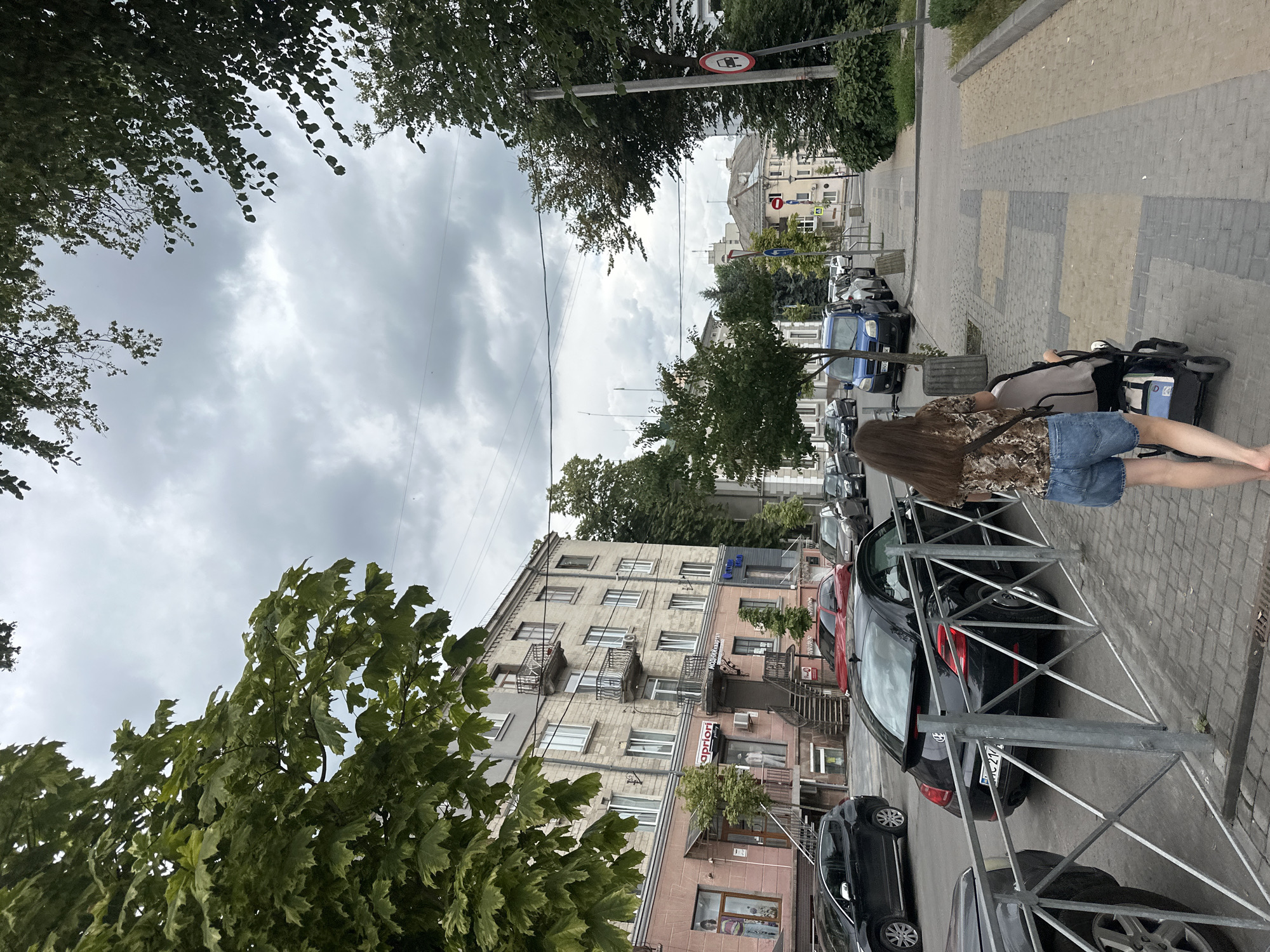 A person is seen from behind pushing a stroller down a sidewalk in Ternopil, Ukraine. The person is wearing a patterned short-sleeved shirt and denim shorts, with a dark strap from a bag across their chest. The stroller appears to be grey and black. They are walking on a paved sidewalk with distinct rectangular sections of grey and tan brickwork. The sidewalk is bordered by a metal fence with a diamond pattern. Beyond the fence, cars are parked along the street, including a black Volkswagen Beetle with a visible license plate. Buildings with multiple stories line the street, featuring balconies and windows. Green trees with dense foliage are present on the left side of the image, and smaller trees and bushes are scattered along the sidewalk and street. The sky is overcast with dark, heavy clouds, suggesting a cloudy day. In the distance, more buildings and trees are visible, and traffic signs, including a "no entry" sign with a red circle and white bar, are present. The overall environment suggests a residential or commercial street in a European city.
