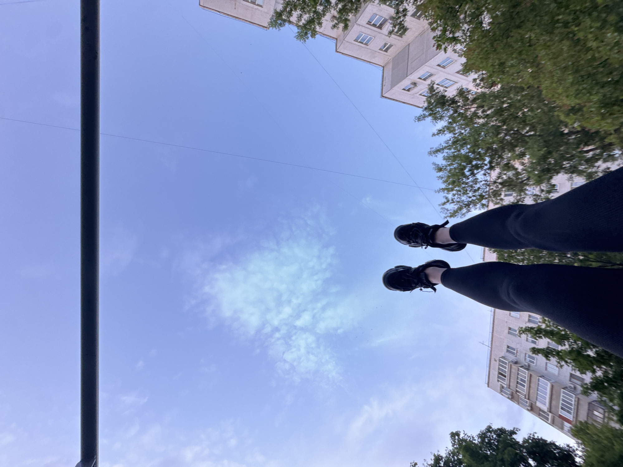 The image is captured from an extreme low-angle perspective, looking upwards towards the sky. In the foreground, a person's legs, clad in dark pants or leggings, and black athletic shoes are prominently featured, extending into the frame from the mid-right and lower-right, elevated against the blue sky. A dark, vertical metal bar stretches from the top to the bottom edge along the left side of the frame, suggesting the person might be hanging from a parallel bar or similar structure, possibly performing an inversion or a physical exercise.
The background consists of a bright, clear blue sky with sparse, wispy white clouds. Thin, dark lines, likely utility wires, traverse the sky horizontally. Parts of multi-story residential buildings, constructed from light-colored concrete or stucco with numerous windows and balconies, are visible in the upper-left, upper-right, and lower-right corners of the frame. Green tree foliage borders these buildings, particularly on the right side.
The bright ambient light indicates it is daytime. The scene is set in what appears to be an urban or suburban residential environment, possibly a park or courtyard equipped with outdoor exercise equipment. The unique perspective captures a dynamic moment of activity. No discernible text is visible.