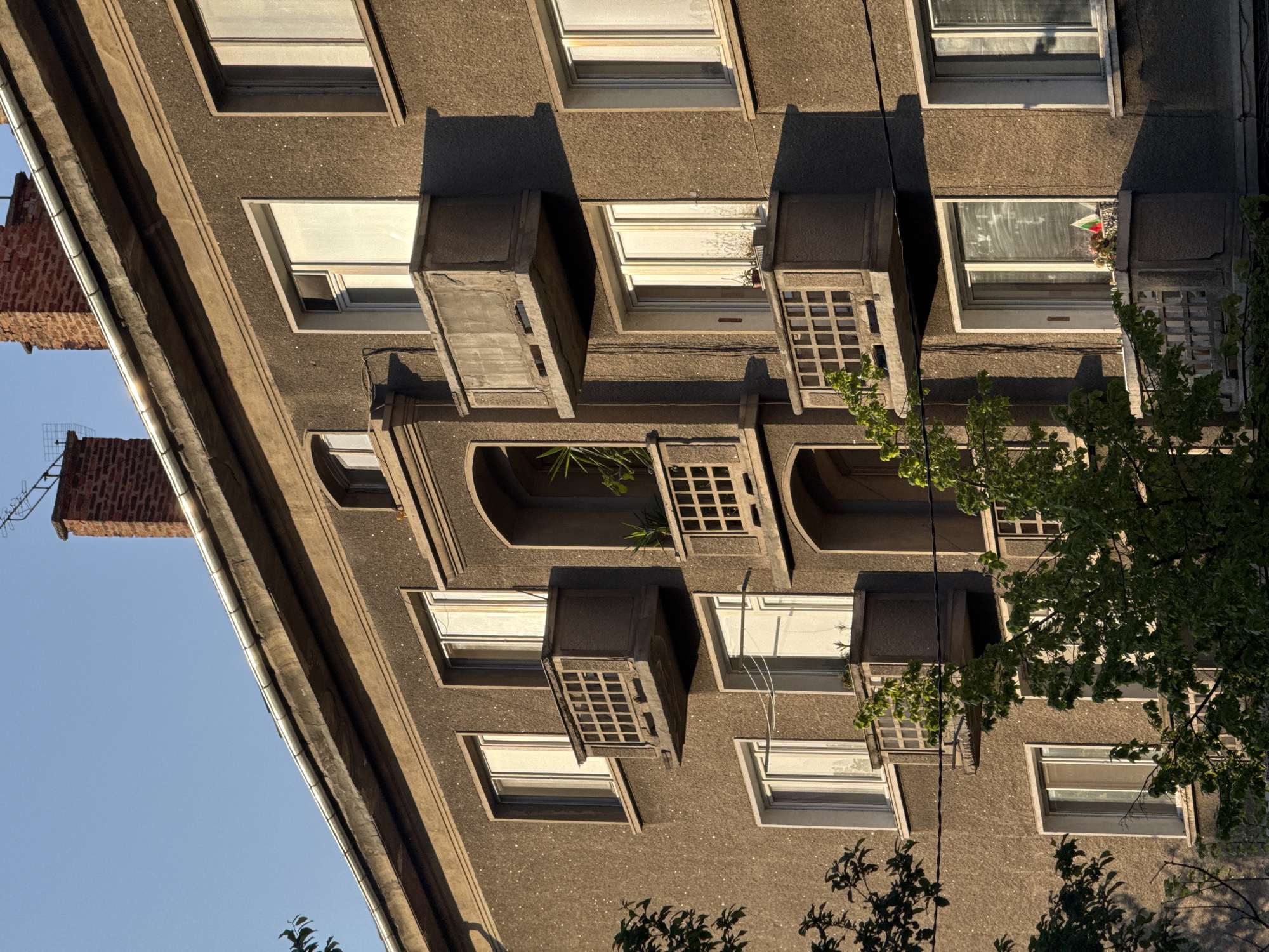 This image captures a close-up view of the exterior of an apartment building, likely in Sofia, Bulgaria, as indicated by the location context and a small flag resembling the Bulgarian flag (white, green, red stripes) visible in one of the window boxes.
The building features a textured, coarse-grained concrete or stucco facade in a muted brownish-grey color. It has multiple rows of rectangular windows with white frames, many reflecting the bright sky. Below several windows are small, projecting concrete balconies or window boxes, some adorned with decorative gridded balustrades. One window box contains sparse foliage, while another on a higher floor holds the small flag and some flowers. A larger, recessed balcony opening with an arched top is visible on an upper floor, containing green plants.
The perspective is upward-looking, showing a steep roofline on the left with a metallic gutter system running along it. Two brick chimneys are visible against a clear, bright blue sky, indicating fair weather. The strong, angular shadows cast by the window frames and balconies suggest the time of day is either morning or late afternoon, with the sun at a low angle. Trees and foliage partially obscure the lower and right portions of the building, casting some shadows and adding greenery to the scene. There are no people visible, nor any discernible text.