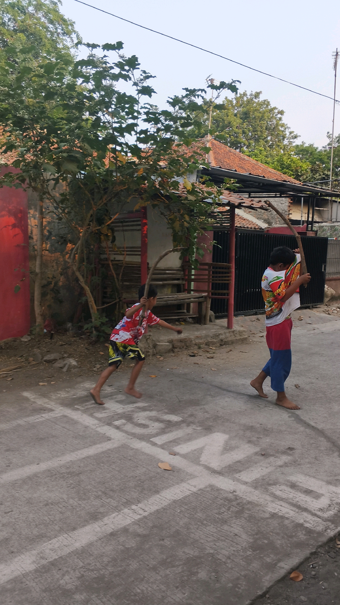 Two barefoot boys are playing on a paved street in what appears to be a residential area of Indramayu, Indonesia. The boy on the left is wearing a red and white patterned t-shirt and patterned shorts, mid-stride as he swings a long wooden stick. The boy on the right is wearing a white t-shirt with a colorful graphic and blue and red shorts, holding a similar wooden stick. White lines are painted on the ground, forming squares, suggesting a game of hopscotch or a similar street game. In the background, there is a red wall, a structure with a tiled roof and wooden beams, and a black metal gate. Trees with green foliage are visible, and the sky is clear. The scene depicts a casual outdoor activity.