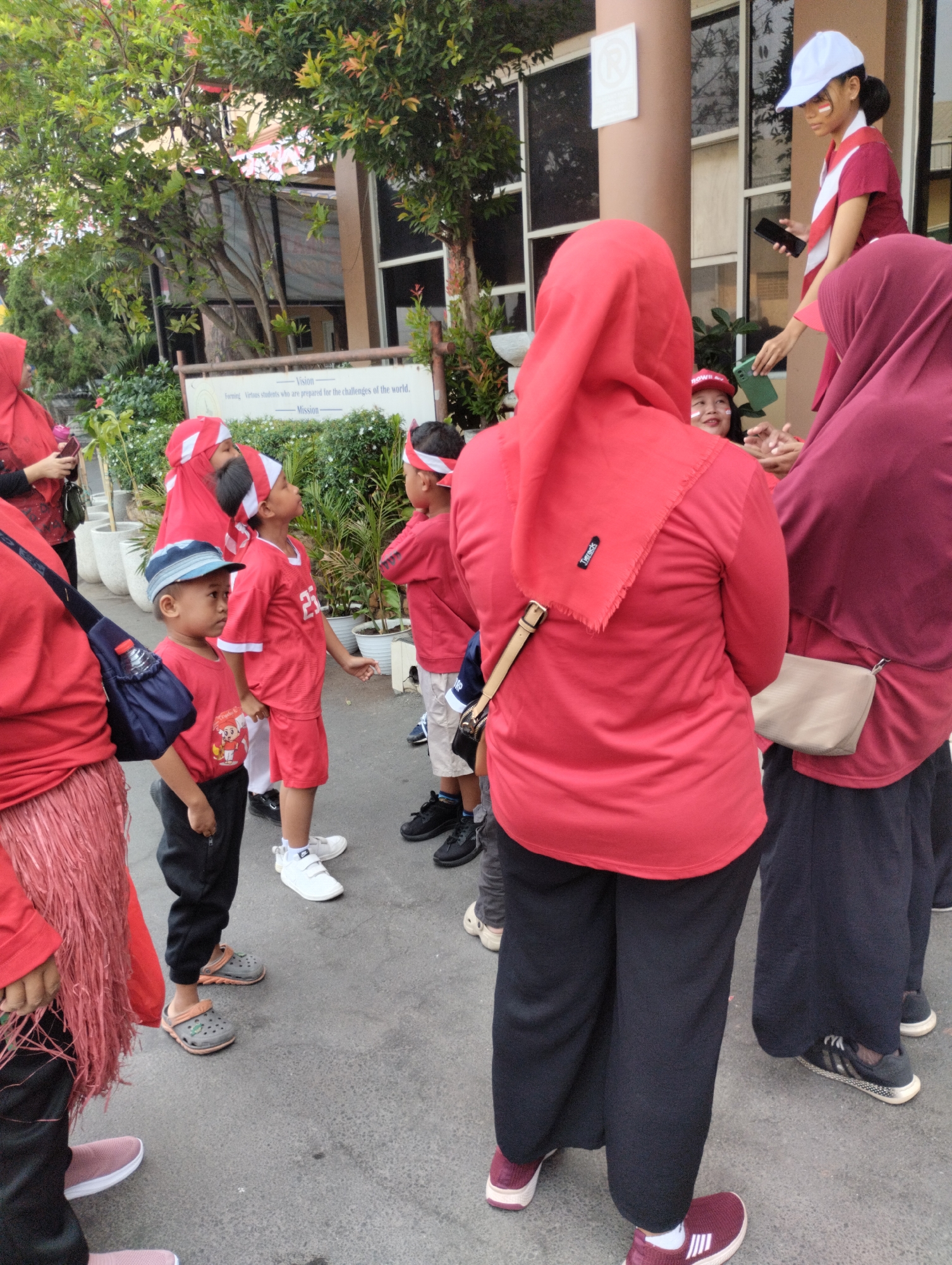 In Bekasi, Indonesia, a group of children and adults wearing red and white attire are gathered outdoors. The children are dressed in matching red shirts and some have red and white headbands. One child wears a blue cap. The adults are also in red tops, with some wearing headscarves. In the background, a building with large windows is visible, along with trees and potted plants. A sign with text is mounted on the building. The ground is paved. The overall scene suggests a cultural event or celebration.