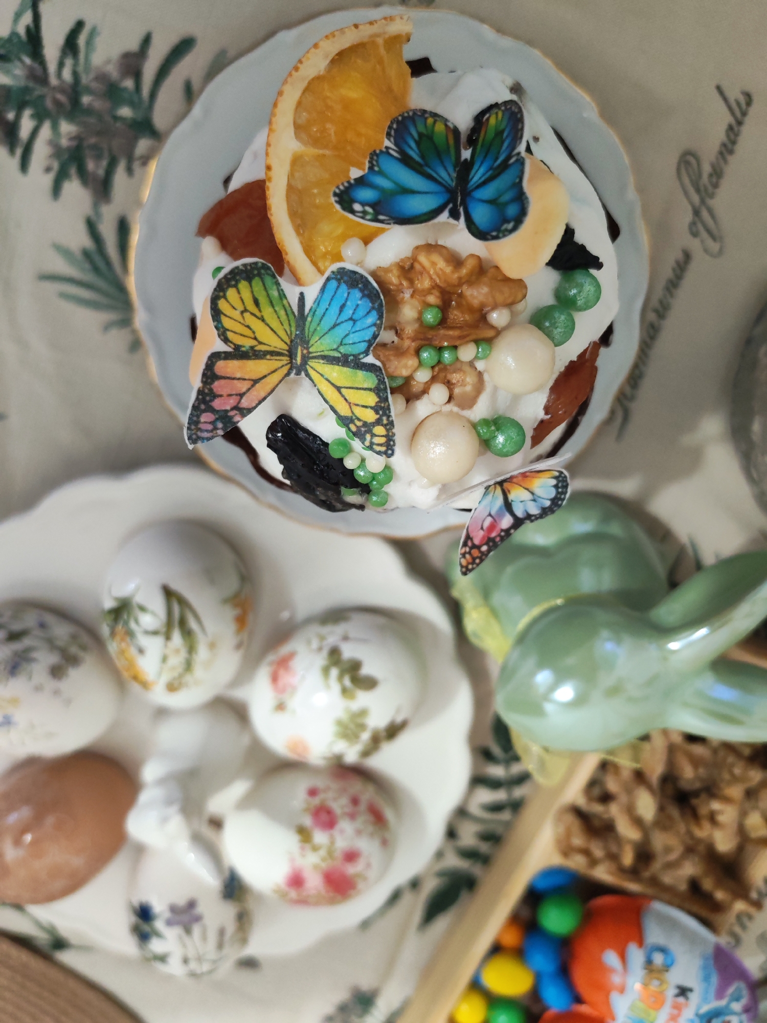 A high-angle shot displays an Easter spread. An Easter cake decorated with white icing, dried fruits, nuts, and butterfly decorations sits atop a light blue plate with a gold rim. Adjacent to it is a plate containing decorated Easter eggs with floral patterns and one brown egg. A green ceramic rabbit figurine is also visible. In the lower right corner, a wooden tray holds walnuts, colorful candies, and a Kinder Surprise egg. The scene is set on a tablecloth with a floral pattern. This scene does not provide enough information to determine if it is located in Izhevsk, Russia.