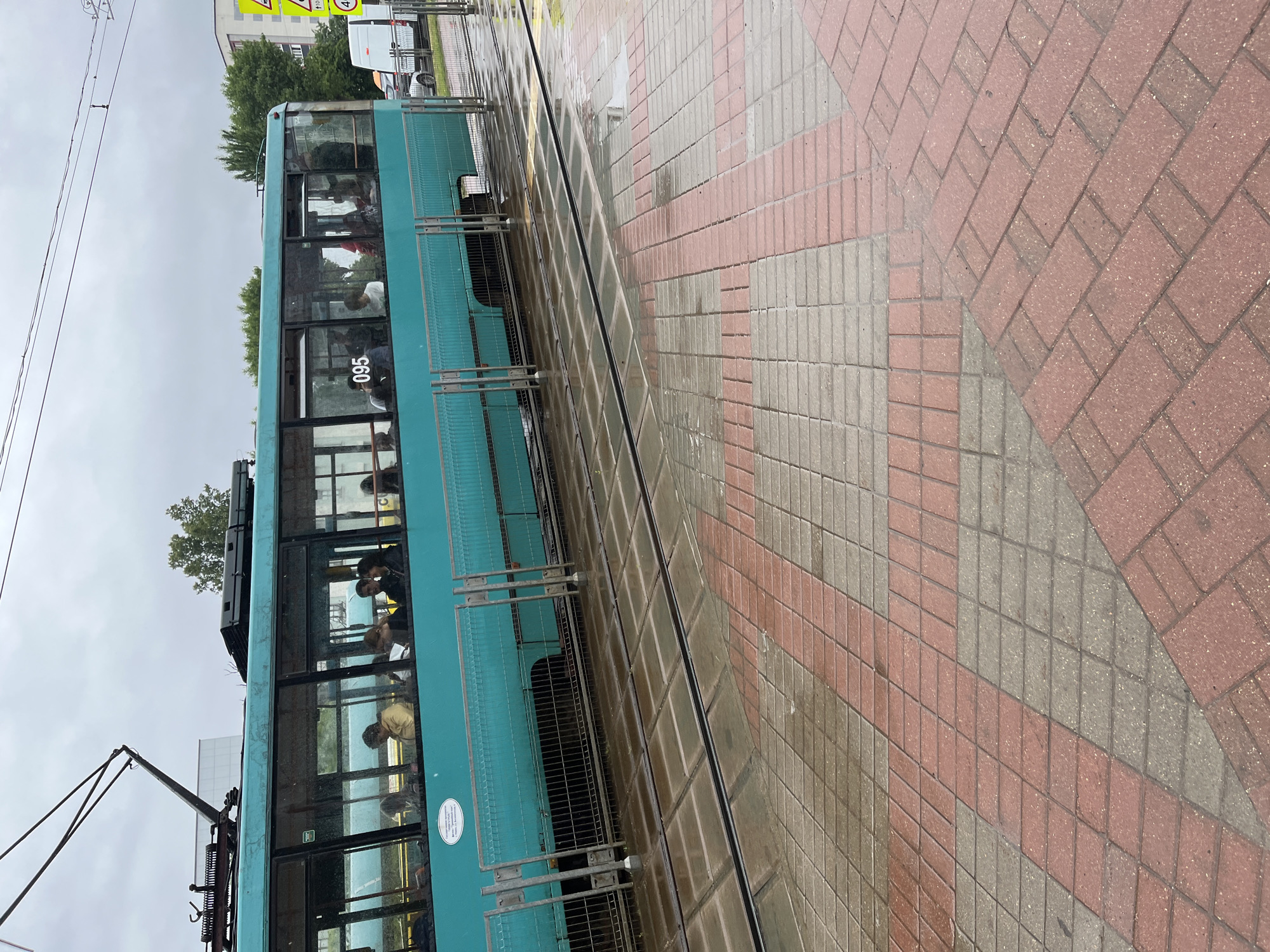 This image depicts a turquoise tram with passengers inside, stopped at a tram stop on a cloudy day. The tram has the number "095" displayed on its side. The tram stop features a wet, red and gray patterned paved area with tram tracks running through it. In the background, there are trees, buildings, and traffic signs, including a yellow diamond-shaped sign with black symbols and a circular sign with the number "40" inside. The scene appears to be an outdoor urban environment, likely a street in Minsk, Belarus, given the distinctive tram design and the visible road signs which are common in Belarus.