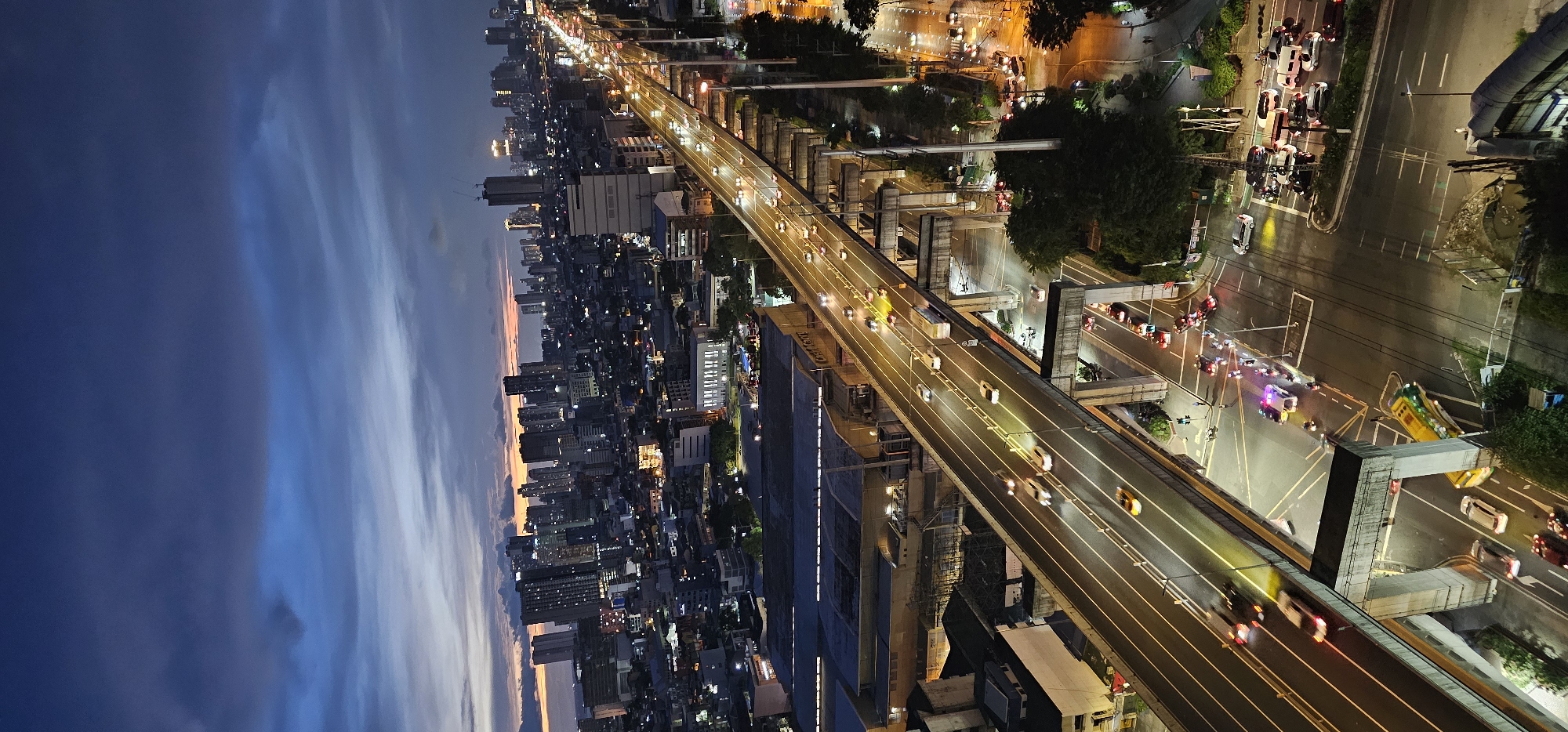 An aerial, long-shot, vertical photograph captures an urban landscape in Makati, Philippines, during dusk. The sky displays a gradient of deep blue, transitioning to lighter blues and subtle hints of orange and pink near the horizon, with dark, streaky clouds across the upper portion of the frame. The cityscape is densely populated with numerous high-rise buildings, many illuminated by internal lights.
A multi-lane elevated highway dominates the mid-ground, carrying a moderate flow of vehicles, their headlights and taillights creating streaks of light. The highway's concrete support structures are clearly visible. Below the highway, a network of roads can be seen, with traffic lights, vehicles, and a yellow bus with green accents visible on the lower road. Several police cars with flashing lights are present at an intersection, suggesting an incident. The scene is well-lit by streetlights and the illuminated buildings, creating a contrast between the dark sky and the vibrant city. The overall impression is one of a busy, dynamic urban environment at the transition from day to night.