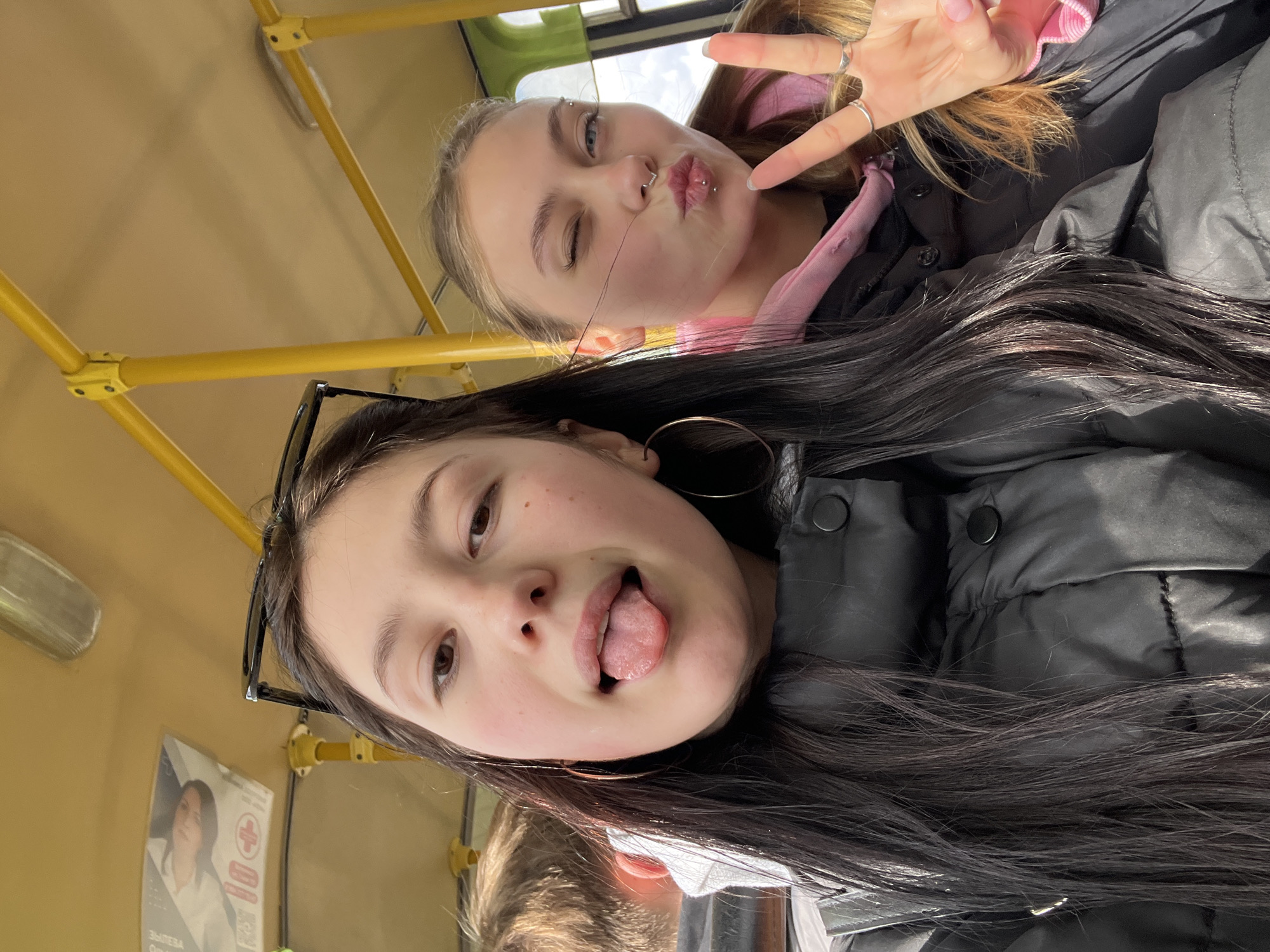 Two young women are captured in a playful selfie, likely inside a bus in Foki, Russia. The image is rotated, showing the subjects from a low-angle perspective.
In the foreground, a young woman with long, dark hair is seen. She wears black sunglasses pushed up onto her head and a black jacket. Her expression is playful, with her tongue sticking out, and she has a silver hoop earring visible. Her cheeks show light blush or freckles. Partially visible below her is another person's head, possibly indicating a third individual or just a tight framing of the foreground subject.
Behind her, another young woman with lighter, possibly blonde or light brown hair, winks and makes a peace sign with her right hand, adorned with silver rings. She has several facial piercings, including philtrum and labret piercings on her lips, and eyebrow piercings. She is wearing a pink top beneath a black jacket.
The setting appears to be a public transport vehicle, strongly suggested by the bright yellow handrails and poles that crisscross the ceiling and walls. A window in the background reveals bright daylight, indicating it is daytime. On the wall, a partially visible poster with Cyrillic text can be seen, clearly showing the word "ЗДОРОВЬЕ" (Zdorovye - Health) and a red cross symbol. The overall mood of the photo is casual and friendly.