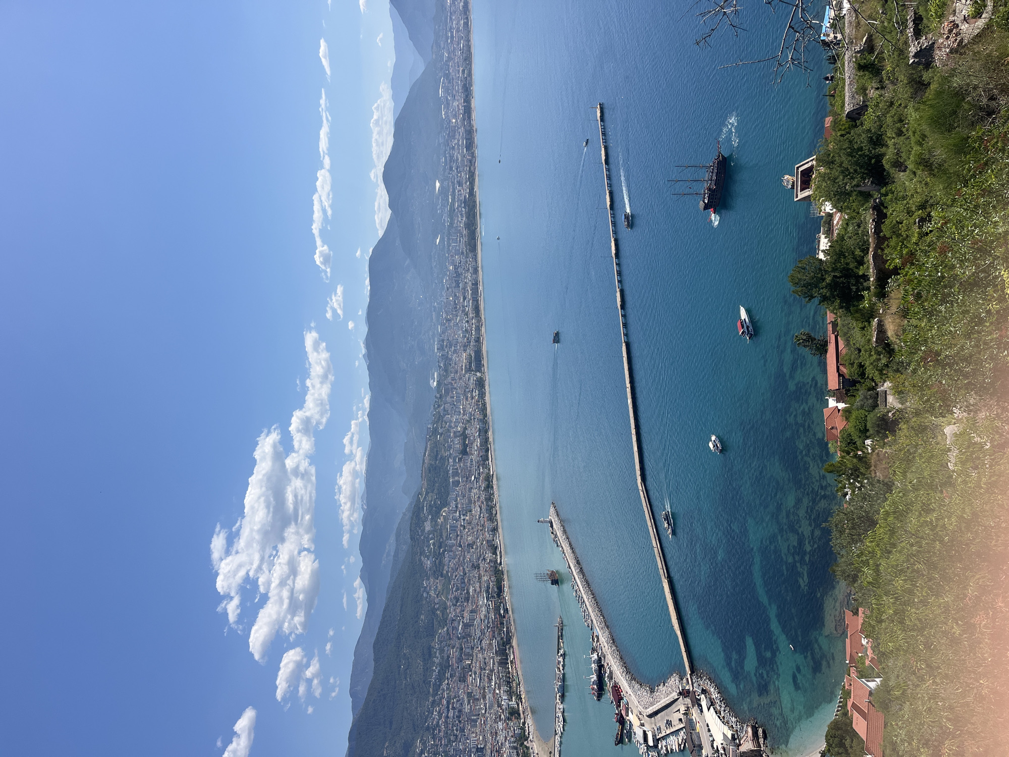 The image captures a high-angle view of Alanya, Turkey, revealing a coastal cityscape nestled between mountains and the Mediterranean Sea. On the left, mountains rise behind a dense urban area that stretches along the coastline. Clear blue skies with scattered clouds are visible above the mountains. The right side of the image is dominated by the sea, with several boats and yachts dotting the water. Two long piers extend into the sea, one with a harbor at its base, filled with docked boats. A large, pirate-style sailing ship is also visible in the sea, moving along the coast. Along the coastline to the right is a stretch of green foliage and buildings. The overall scene depicts a blend of natural landscapes and urban development characteristic of the coastal city of Alanya.