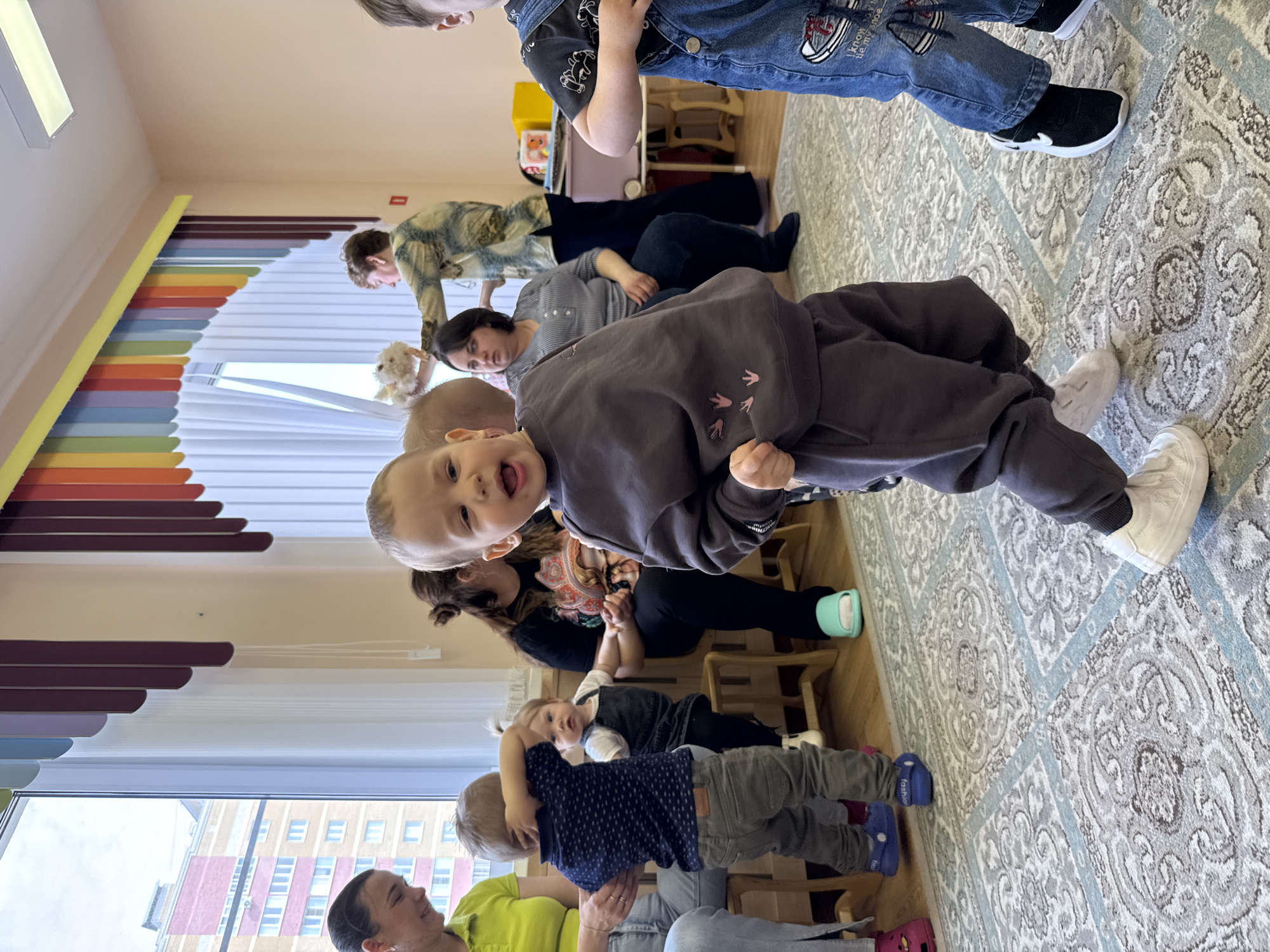 The image shows a group of people, mostly young children, inside a room. The location is presumably Rusanovo, Russia.
There are multiple adults interspersed among the children, suggesting a caregiver or educational setting. The children are seated or standing close to the adults.
The room has a carpet with a patterned design. Near the window are colorful decorations above it. Outside the window, an apartment building can be seen.