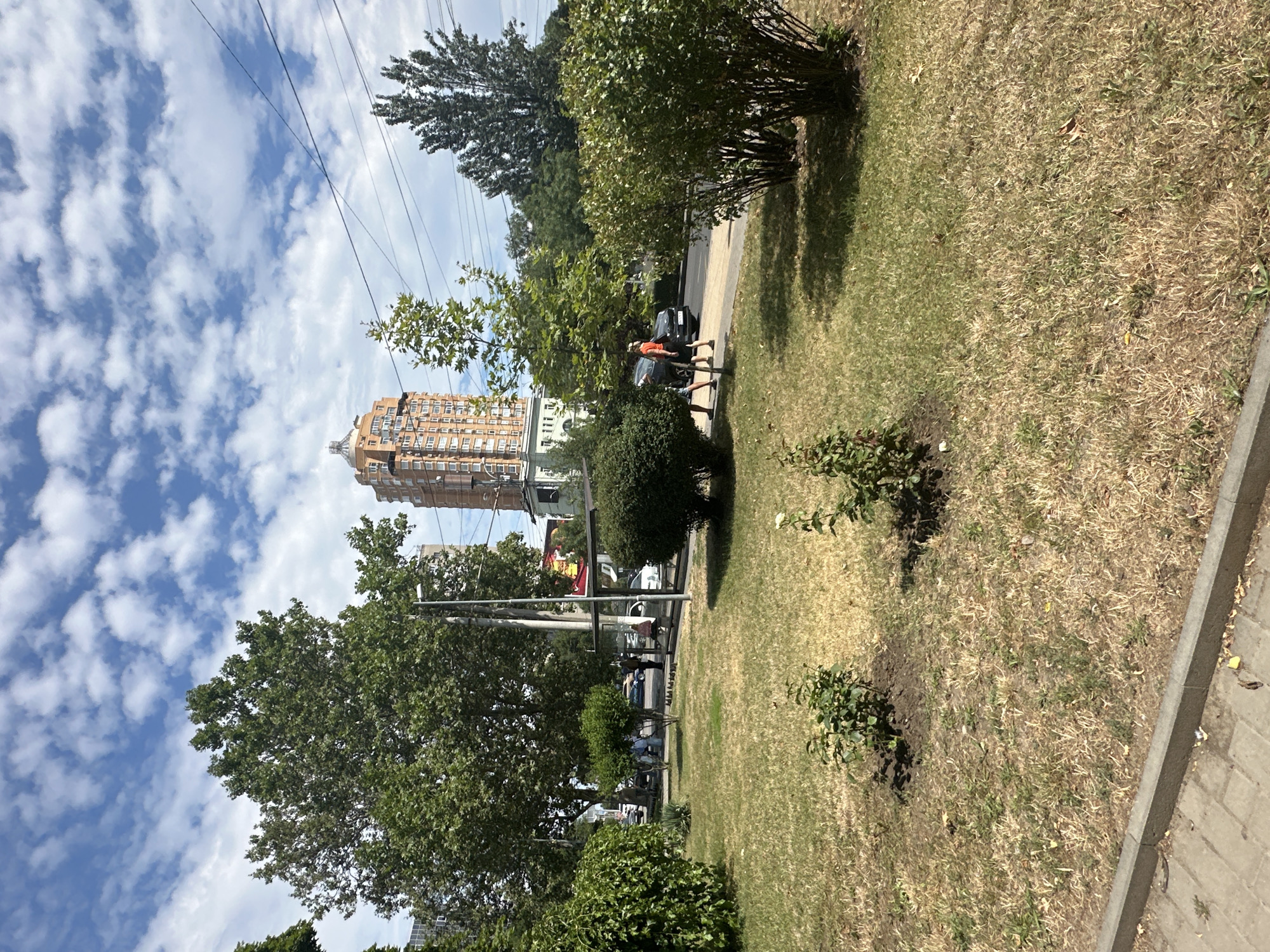 In Odesa, Ukraine, a street scene unfolds under a partly cloudy sky. In the foreground, a patch of dry, sun-baked grass is bordered by a sidewalk made of grey paving stones. A small, leafy bush grows in the grass on the left, and another, slightly larger one, is visible on the right.
Behind the grass, a paved area features a bus stop shelter with a few individuals visible within. To the left of the bus stop, cars are parked along the street. A tall, multi-story building with a distinctive orange facade and a rounded top dominates the background. Several mature trees with dense green foliage are scattered throughout the scene, some appearing to be deciduous. Power lines crisscross the sky above the trees and buildings.
A dark-colored car is traveling on the road in front of the bus stop, and two people are walking on the sidewalk beside it. One person wears an orange shirt and dark shorts, while the other wears dark shorts and a lighter top. The overall impression is a typical urban environment during daylight hours.