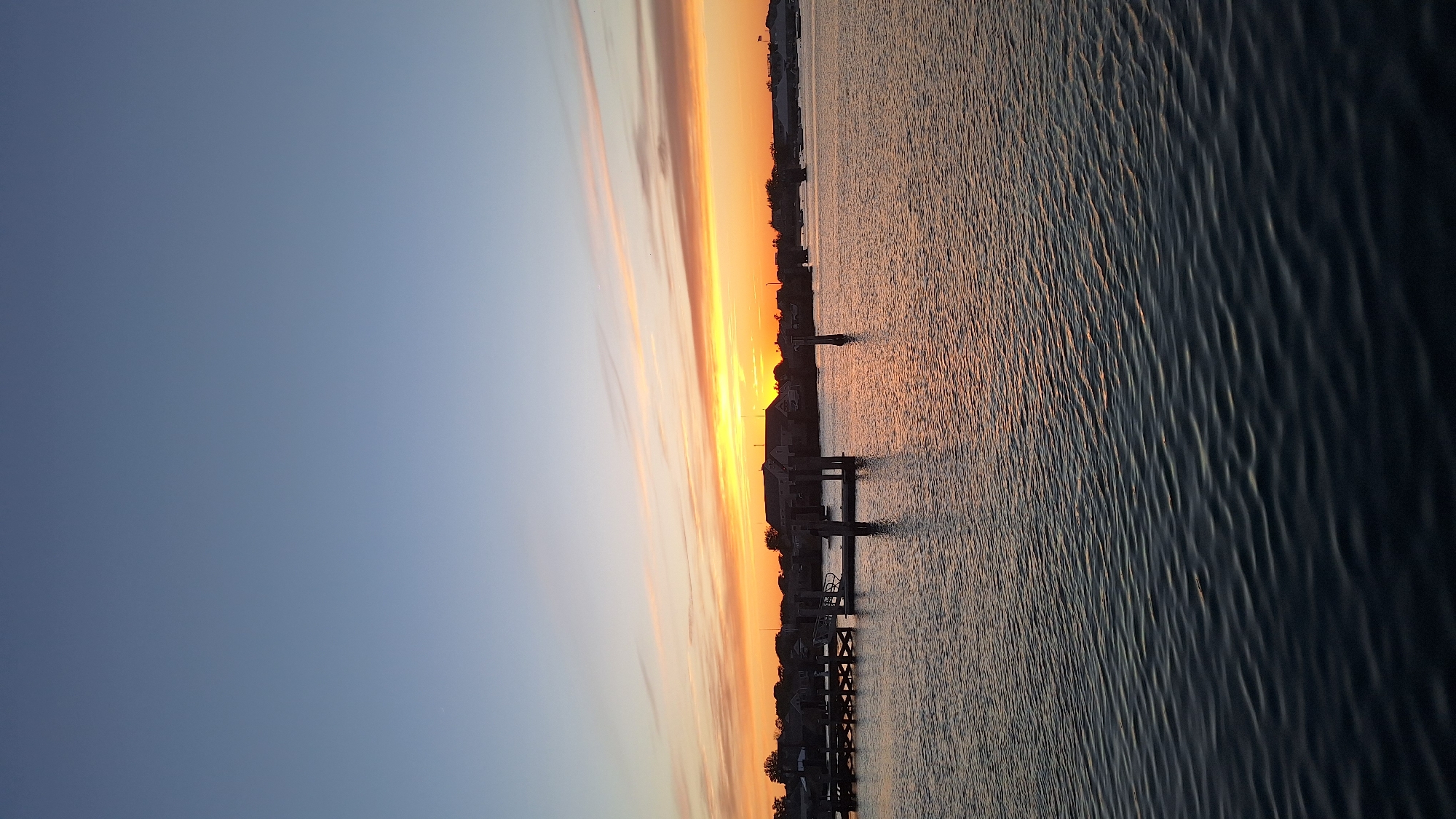 The image depicts a serene waterfront scene at dusk on Nantucket, Massachusetts, United States of America. The sky transitions from a pale blue at the top to a vibrant spectrum of orange and yellow near the horizon, indicative of a sunset. Wispy clouds are scattered across the sky, catching the fading sunlight.
In the foreground, a body of calm water reflects the colors of the sky and the surrounding structures. The water's surface is rippled, creating subtle patterns.
Along the shoreline, a wooden dock with pilings is visible, extending into the water. This dock appears to be a mooring area for boats, though no vessels are prominently featured in the immediate foreground. Further back, buildings with dark roofs and windows are silhouetted against the colorful sky, suggesting a residential or commercial area bordering the water. Trees and vegetation are also present along the coastline.
The overall atmosphere is tranquil and peaceful, capturing the quiet beauty of a coastal evening. There are no people or animals visible in the image. The scene is static, focusing on the natural beauty of the sunset and the architecture of the waterfront.