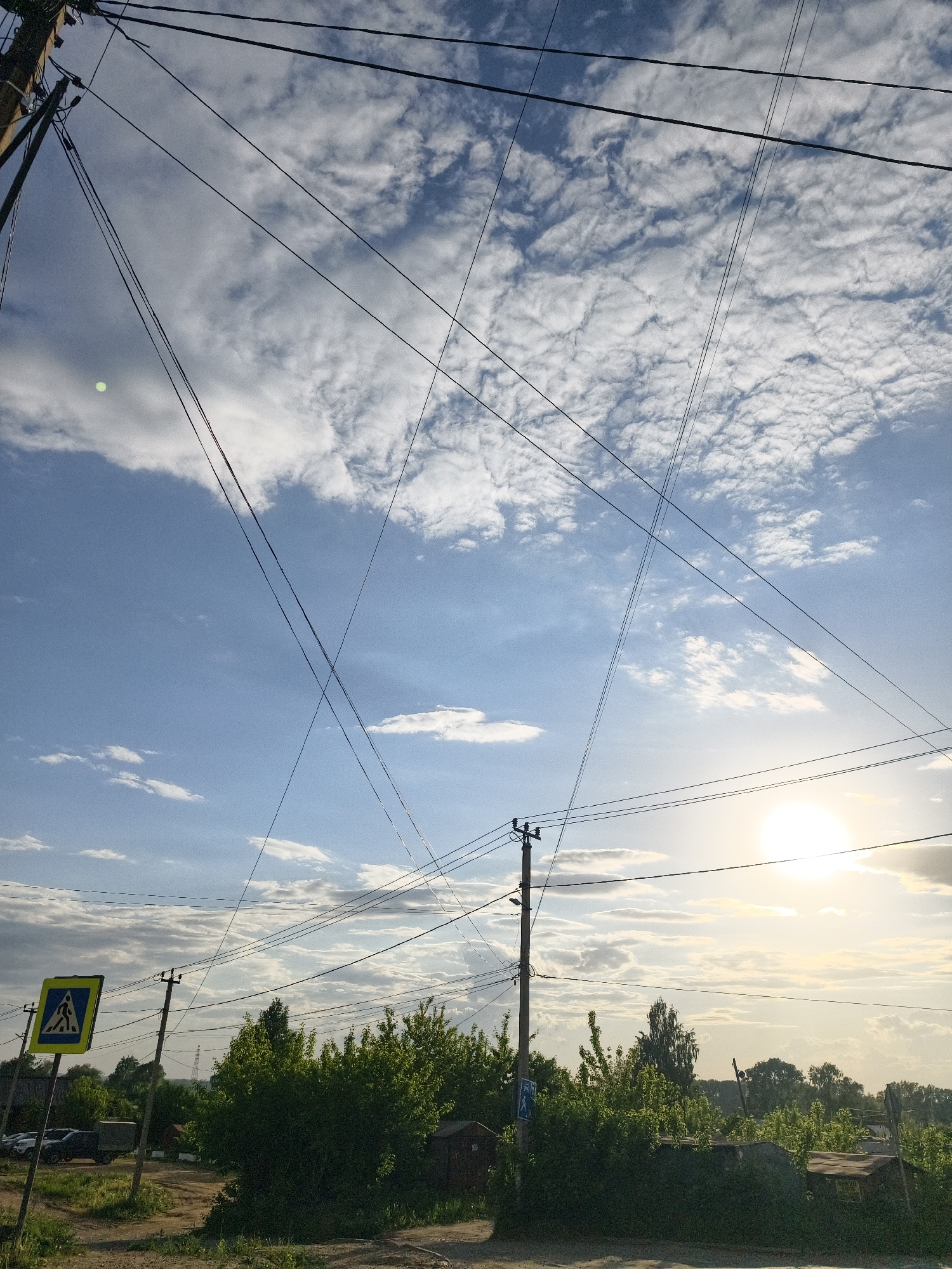 The image captures a street view in what appears to be a residential area, possibly in Chaikovsky, Russia, as specified. Power lines crisscross the sky above. The sky is blue with patchy white clouds. The sun is partially obscured by clouds and atmospheric haze, creating a bright spot in the sky.
At ground level, there are green bushes and trees. A pedestrian crossing sign is visible on the left side of the image. Cars and small buildings can be seen in the background. The road appears unpaved and dirt.