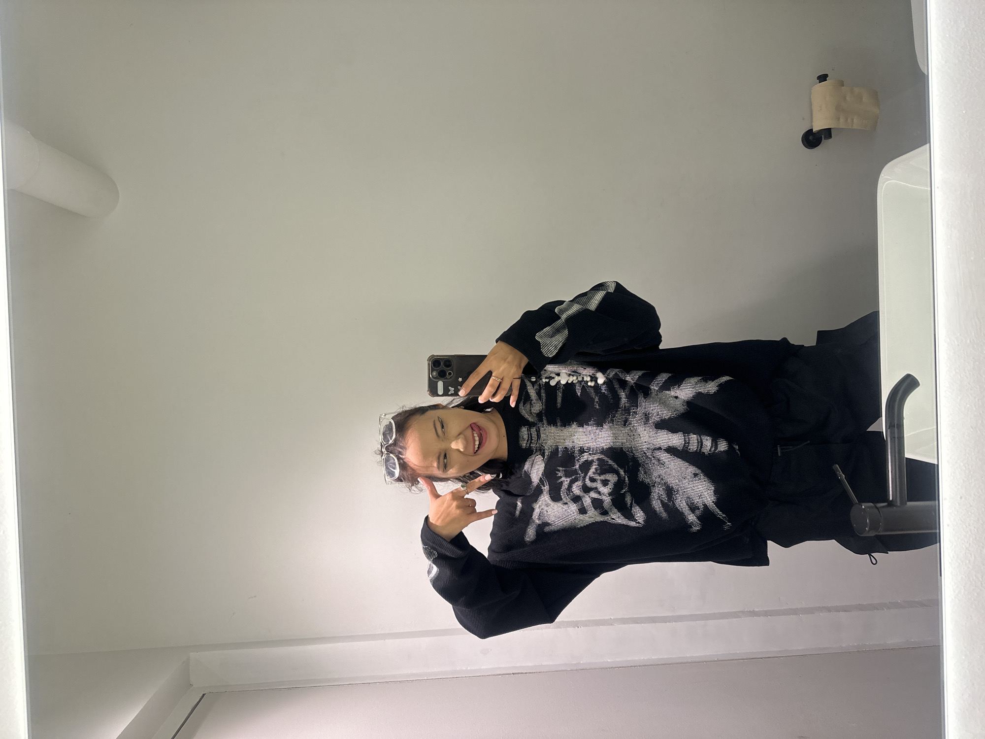 A young woman with dark hair and sunglasses perched on her head takes a mirror selfie in a bathroom. She is wearing a black long-sleeved sweater with a white skeleton print and black pants. She sticks her tongue out and makes a rock-and-roll hand gesture. In the background, a toilet paper holder with a roll of toilet paper is mounted on the wall. The sink and faucet are visible in the foreground. The scene takes place in a modern bathroom with white walls and a large mirror.