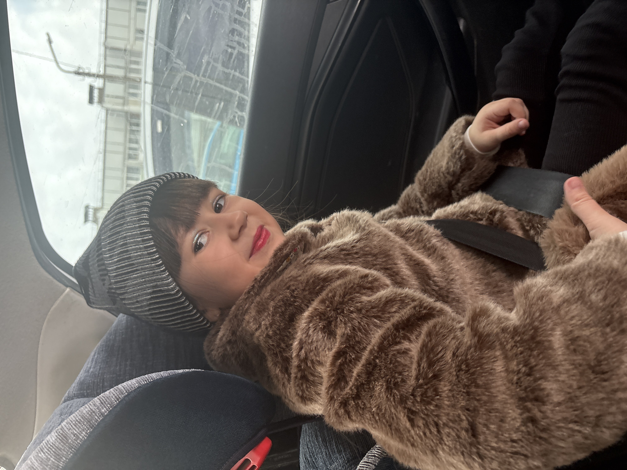A small child, likely a girl, is sitting in a car seat in a vehicle. She's wearing a striped knit hat and a brown faux fur coat. Her lips are visibly red. She is strapped into her seat with a black seatbelt. Another person, probably an adult, is sitting next to her. Outside the car window, blurred buildings are visible. Based on the window reflection of tall buildings, the location appears to be an urban environment, likely Chaikovsky, Russia.