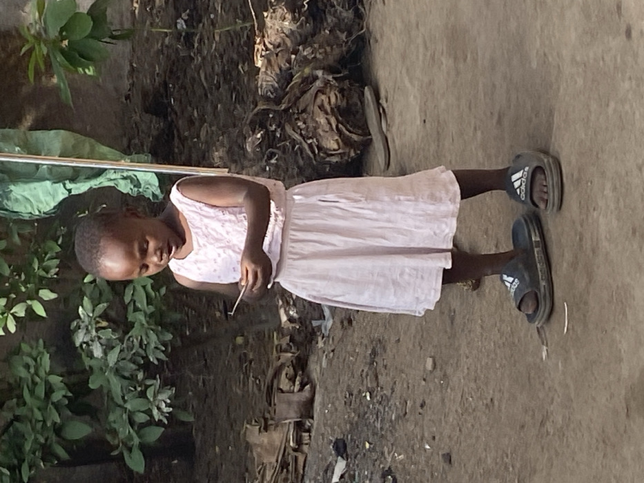 An outdoor, eye-level photograph captured in Udu, Nigeria, depicts a young Black child standing on bare earth. The child, with short dark hair, wears a light pink sleeveless top with a textured pattern and a matching plain light pink skirt. Oversized dark grey "adidas" brand slide sandals are on her feet. She is looking downwards with a focused expression, her mouth slightly ajar, as she manipulates a small, thin, light-colored object held between both hands at chest height. The background consists of green foliage on the left and right, with a notable green fabric draped vertically on the far right. The ground is irregular dirt, with scattered organic debris, including what appears to be a single brown boot lying on its side in the mid-left background. The scene is illuminated by natural daylight.