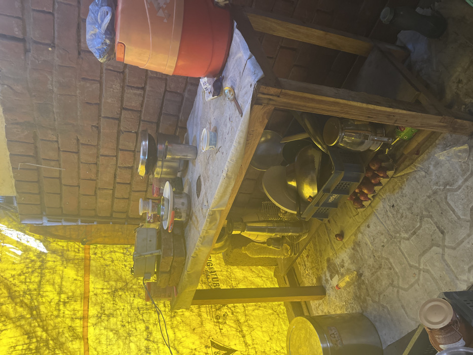 The image shows a wooden table with various items on it, including a metal container, a blue container, a plastic bottle, and a metal tea pot. The table is located in an outdoor area with a brick wall to the right. Behind the table is a large yellow tarp that is stretched across the area. The location of the scene is Sahibzada Ajit Singh Nagar, India, based on the provided coordinates. The environment is dusty and worn. There is a stainless steel water jug to the right of the table. There are also small red onions on the ground, indicating that this may be a food preparation area. The image is well-lit, and there are no people present.