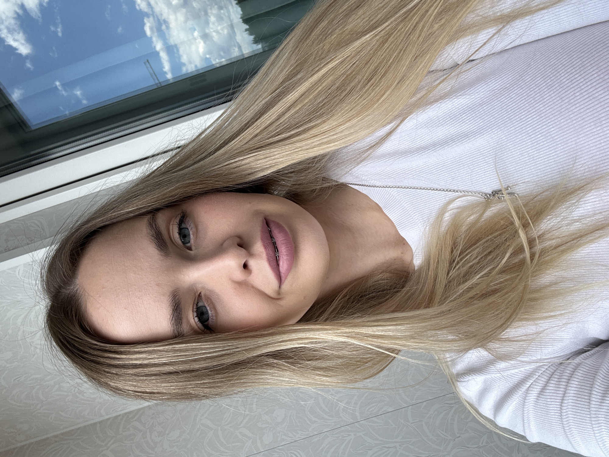 A young woman with long blonde hair is pictured indoors, possibly in the city of Chaikovsky, Russia. Her hair is parted to the side and falls around her face and shoulders. She has blue eyes and is wearing clear braces on her teeth. She is wearing a white, long-sleeved shirt. In the background, a window reflects a bright blue sky with clouds. The floor has a patterned design. A silver chain necklace is visible.