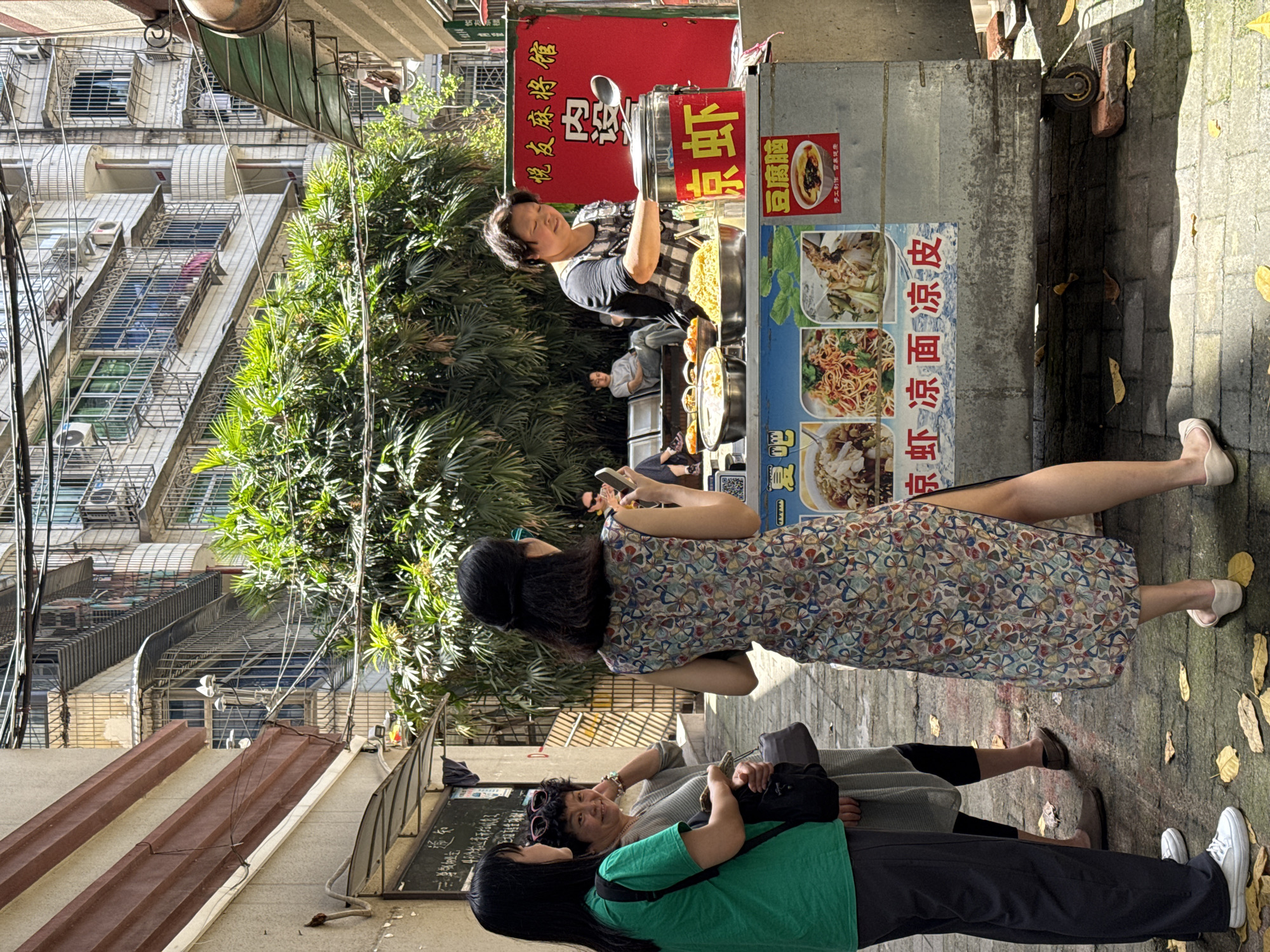 Here is a detailed description of the image:
**Setting:** The image captures a street scene in what appears to be a residential area in the Yuzhong District of China. Tall buildings with metal balconies and air conditioning units line one side of the narrow street. Green trees add a touch of nature amidst the urban setting. The ground is paved with stone blocks.
**People:**
1. **Vendor:** A middle-aged person, likely the vendor, is seen behind a food stall.
2. **Customers:** Three women are standing near the stall. One woman in a floral dress holds a phone in front of the stall. One is wearing a green shirt and black pants. All three are interacting with the vendor.
**Objects:**
1. **Food stall:** A metal food stall is set up on the street. It has signs with Chinese characters and images of food dishes, indicating the kind of food being sold.
2. **Signage:** Red signs are seen on the food stall.
3. **Leaves:** Scattered leaves cover the ground.
**Action:**
1. The vendor is attending the food stall.
2. One woman is holding a phone near the stall.
**Environment:**
The setting includes high-rise buildings, greenery, and a narrow street, suggesting a lively yet confined urban environment.