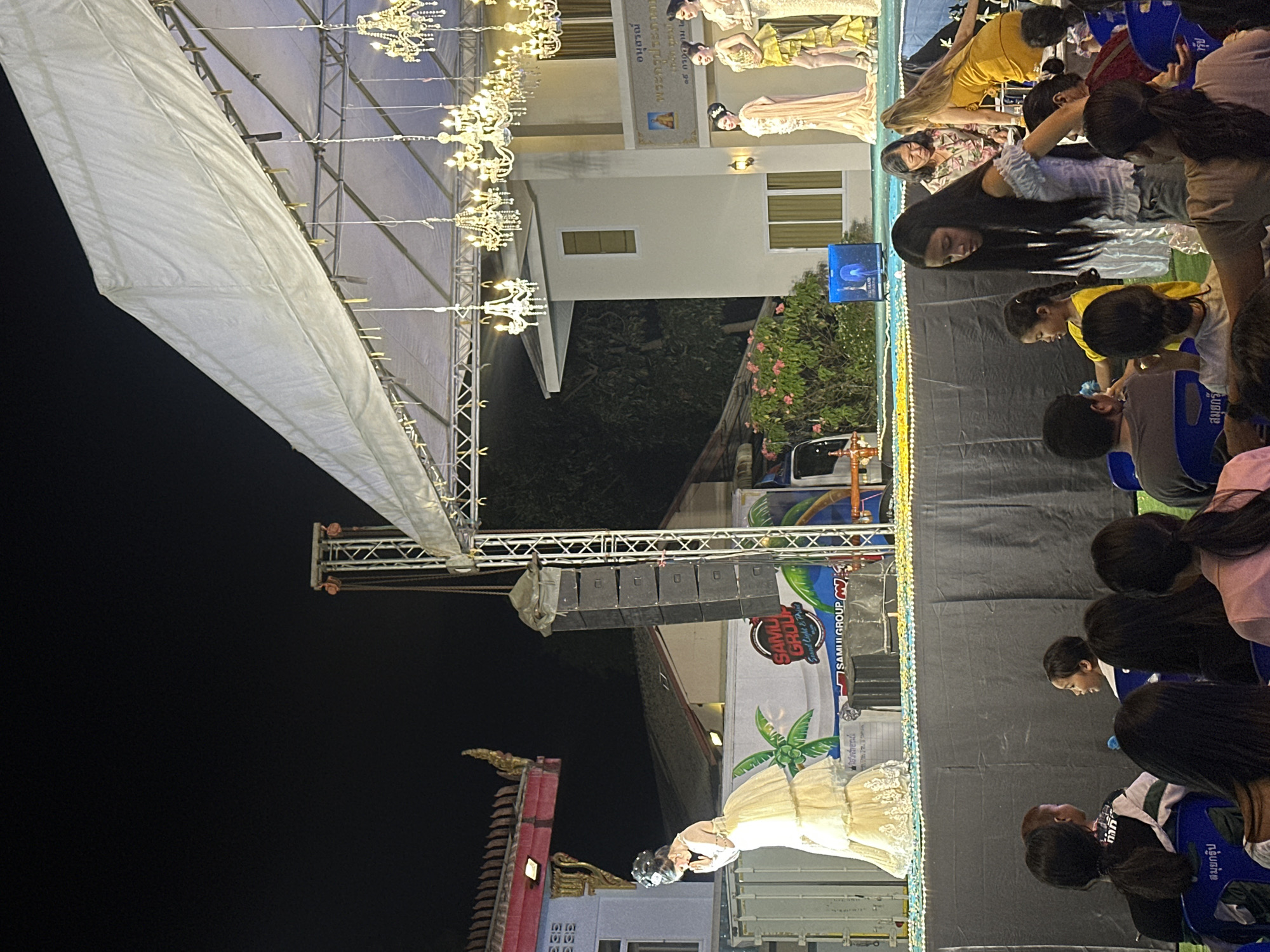 Here is a detailed description of the image:
**General Description:**
The image shows a staged performance or event, likely in an outdoor setting during the evening. There is a covered stage with chandeliers hanging overhead, and a backdrop curtain behind the performers. A crowd is seated facing the stage.
**People:**
* There are multiple people visible in the image.
* Several individuals are performing on stage wearing traditional Thai clothing, with at least 3 visible.
* A crowd of people is seated in front of the stage. Their actions consist primarily of watching the performance.
* The seated audience members are mostly facing the stage.
**Subjects/Objects:**
* A large white canopy structure above the stage, likely providing cover.
* Multiple chandeliers hanging from the stage structure.
* A stage backdrop that appears to be black.
* Speakers and other equipment are positioned on either side of the stage.
* Seating arrangement with chairs for the audience.
* A vehicle and signage is visible behind the performance area
**Actions/Interactions:**
* Performers are on stage.
* The audience is watching the performance.
**Environment:**
* The image appears to be taken in the evening or at night.
* The location appears to be บ้านบางรักษ์, Thailand, based on the OCR text, including the text สมุยกรุ๊ป, commonly associated with Samui Group which provides support for events and lighting within the district.