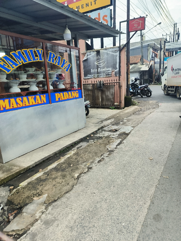The image shows a street scene in the city of Ngamprah, Indonesia (latitude: -6.867, longitude: 107.518). The scene is dominated by a street with a concrete surface. The street is cracked and uneven, with patches of asphalt and dirt. A small food stall with a sign that reads "Family Raya Masakan Padang" is located on the left side of the street. The stall appears to sell Padang cuisine. There is also a sign advertising "Soto Bandung" on the wall of a building next to the stall. A motorcycle is parked in front of the stall, and a truck is parked further down the street. The truck is hauling a large container, the signage on which indicates that it is "Central Cargo." The truck is parked on a slightly wider street, with the road joining the narrow street in front of the food stall. The street is partially obscured by the stall and the building to its right. In the distance, the buildings appear to be mostly residential. The sky is cloudy, and the weather appears overcast. There are no people visible in the image.