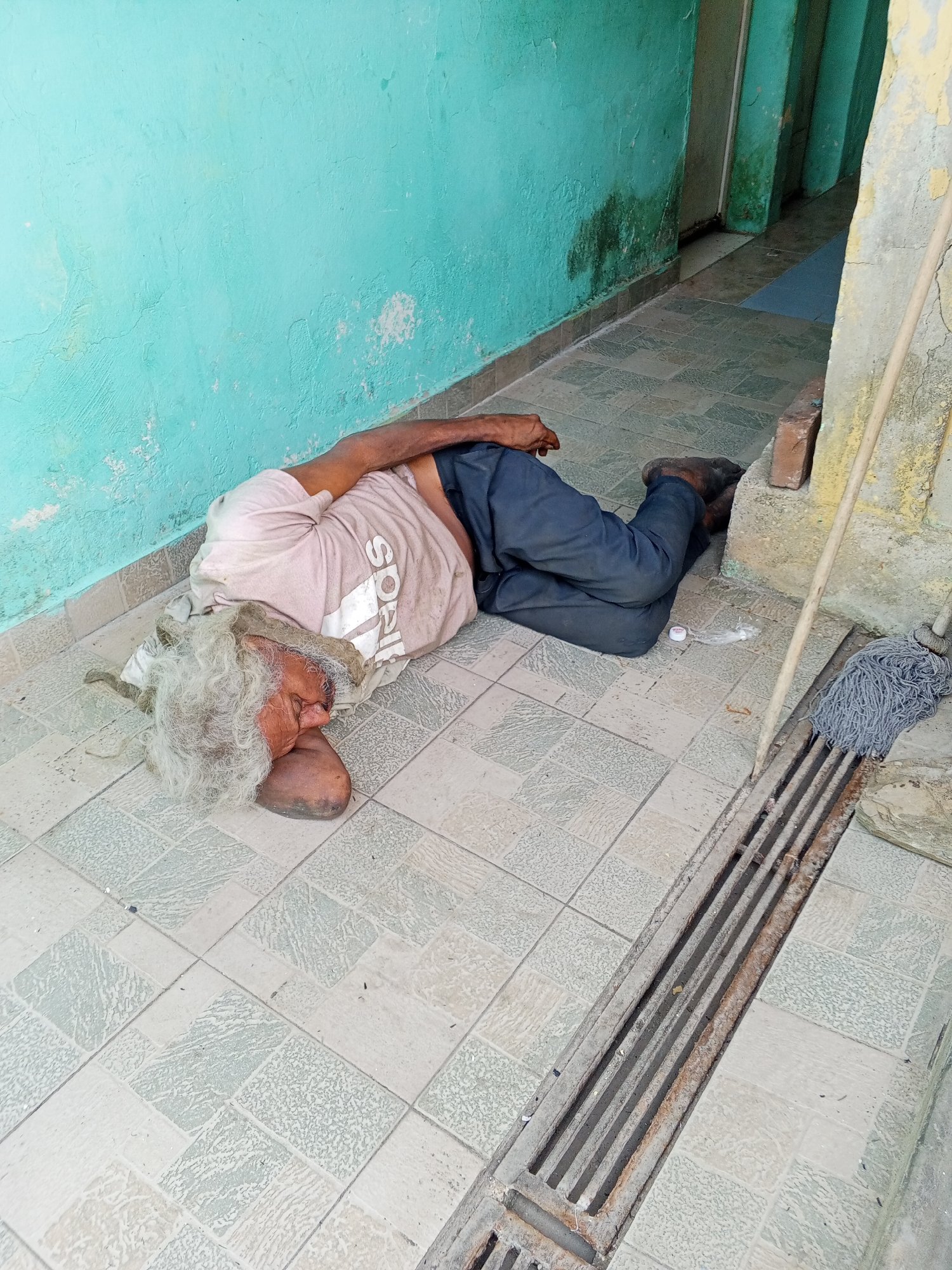 A man is lying on a tiled floor next to a turquoise wall and a drain. The man has long, gray hair and is wearing a light pink shirt and dark pants. His head is resting on his arm. A mop leans against the wall near the drain. The location is likely in Tinaquillo, Venezuela.