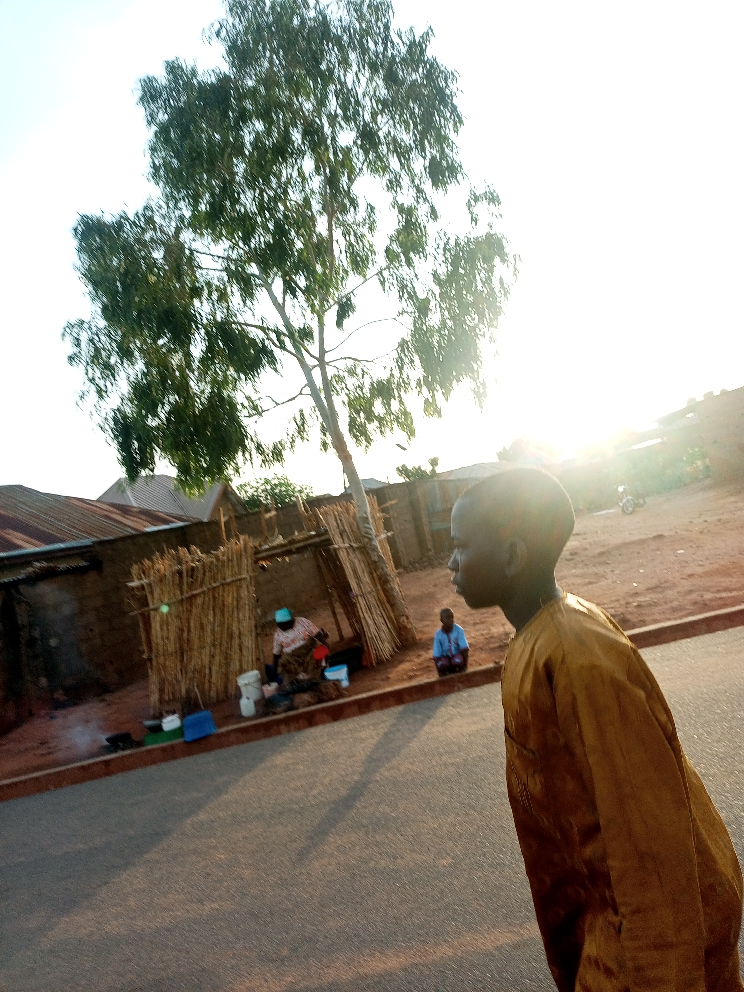 The image depicts a daytime scene in what appears to be a rural or less developed area. In the foreground, a person wearing a mustard-colored robe is walking along a paved road. Their facial features are partially obscured by the sun's glare, but they seem to have a dark complexion and a shaved head.
In the background, there is a simple dwelling with a tin roof and a structure made of vertical sticks. A woman wearing a patterned dress and a light blue hat is seated nearby, possibly preparing food, as there are pots and other cooking implements around her. A young boy in a blue shirt is sitting on the ground a little further away.
Behind the structures, there is a tree and a dirt road leading to other buildings in the distance. The lighting is bright, with the sun shining directly into the camera, causing a bright glare. The overall impression is that of a simple, perhaps impoverished, community. The exact location and city are not discernible from the image.