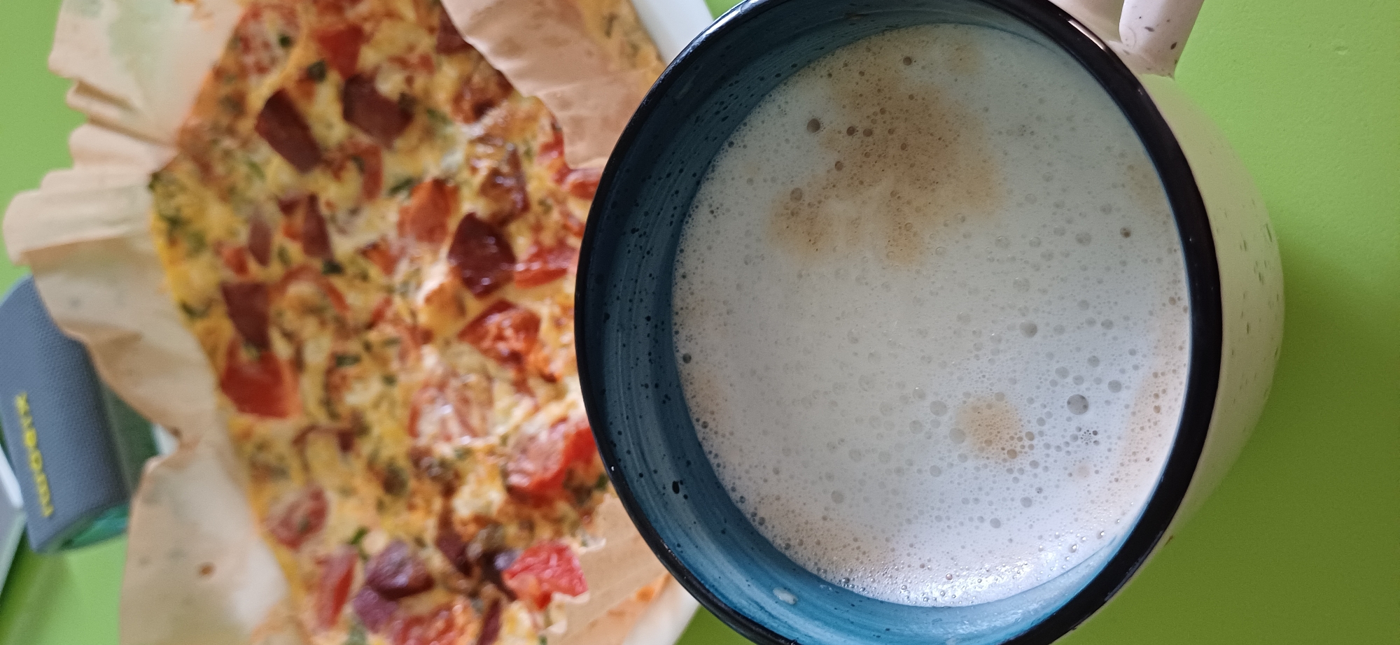 The image shows a close-up view of a cup of coffee with froth in the foreground. The coffee is in a blue and white mug. Behind the mug, a dish of food, possibly a frittata or quiche with tomatoes and sausage, is visible, placed in a paper liner. In the background, a blurred grey object with yellow lettering is present. The entire scene is set against a bright green surface. There are no people or identifiable city landmarks in the image.