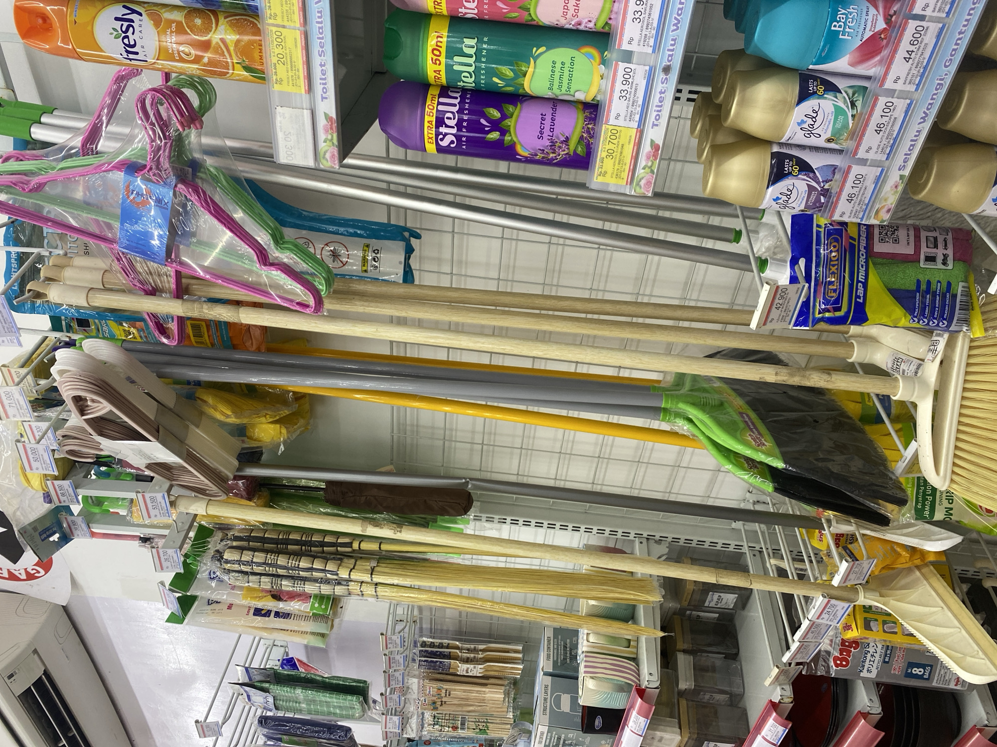 The image shows a retail store aisle in Bantaeng, Indonesia, stocked with household cleaning supplies. The shelves are filled with brooms, mops, dustpans, air fresheners, and other cleaning tools. Products are displayed in packaging with price tags in Indonesian Rupiah (Rp). The lighting appears to be fluorescent, typical of a supermarket or minimarket. There are no people visible in the image. The store environment suggests a well-organized display of cleaning products for sale.